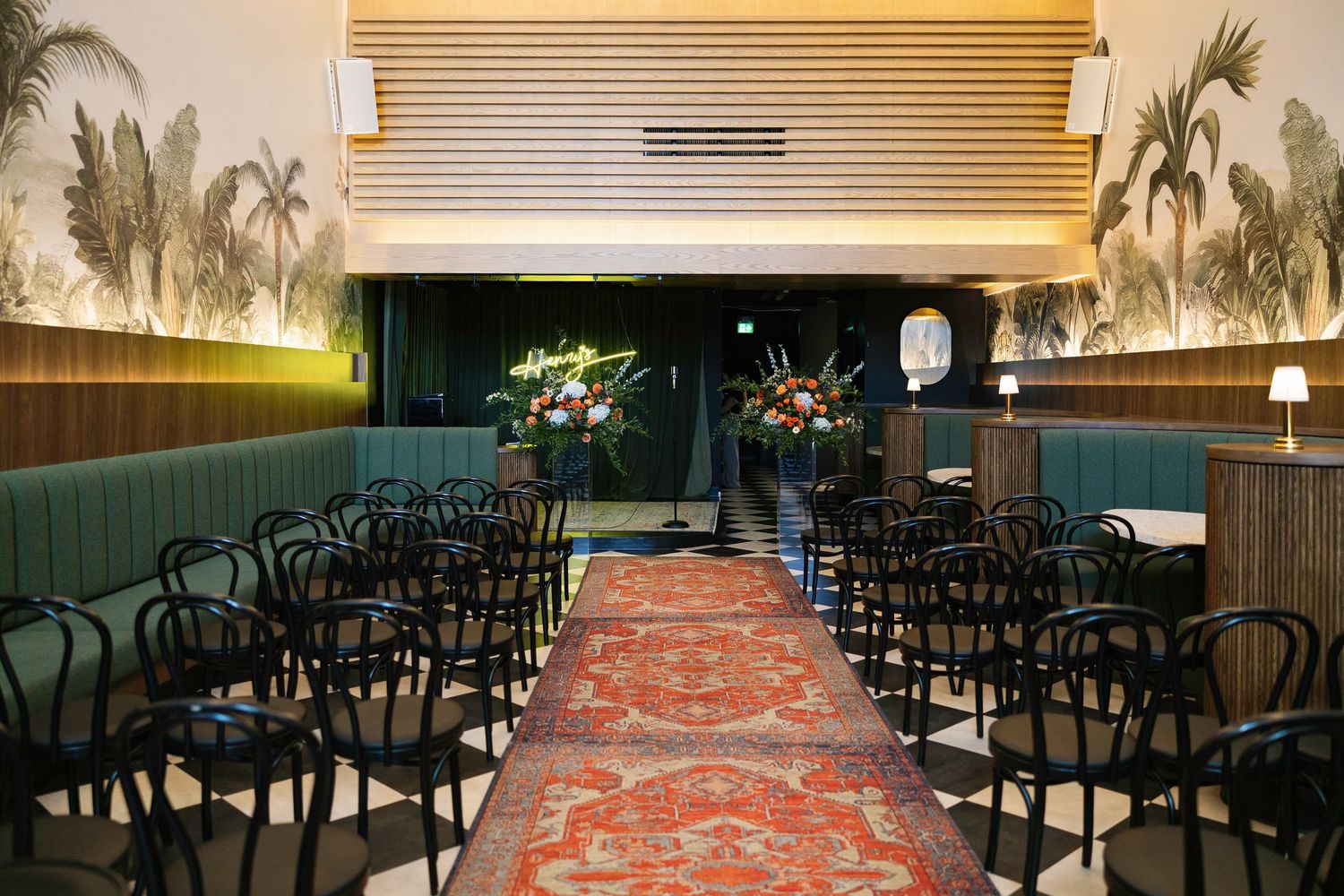 Elegant wedding ceremony venue with black chairs, red patterned aisle runner, floral arrangements, and tropical wall murals.