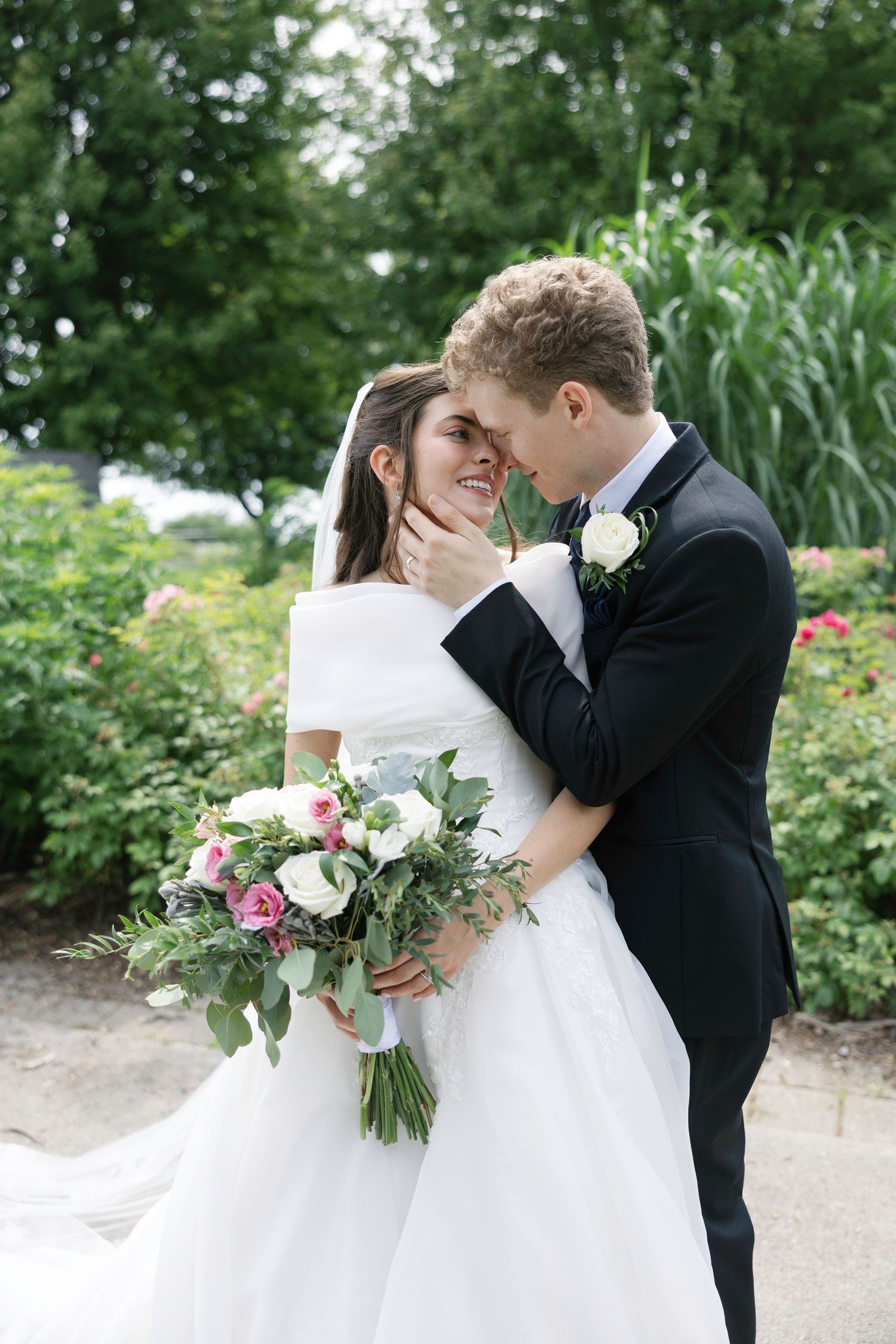 A bride and groom share a romantic moment in a lush garden, she holds a pink and white bouquet in a flowing white gown.