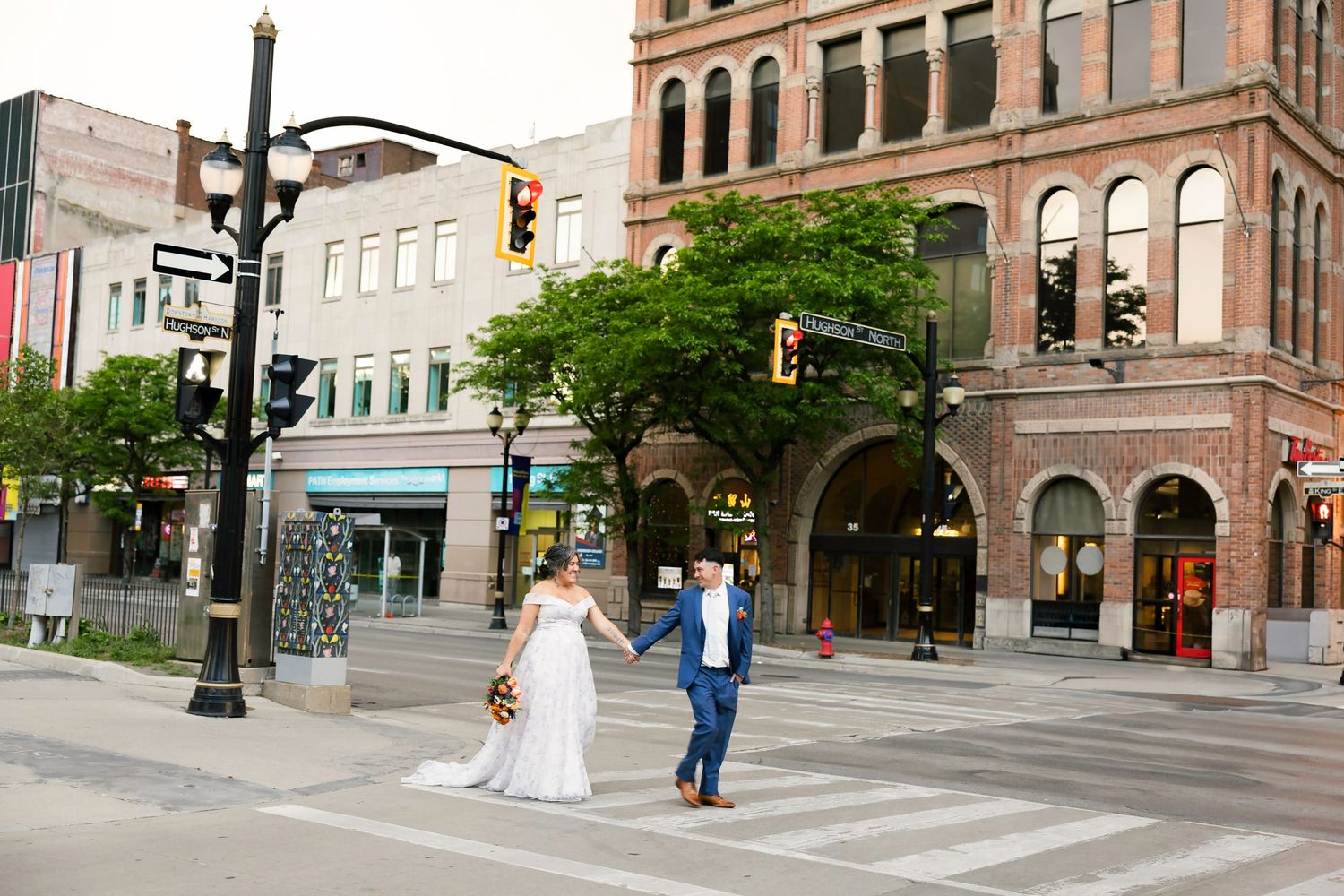 Bride and groom holding hands crossing a city street intersection in downtown, with historic brick buildings in the background.
