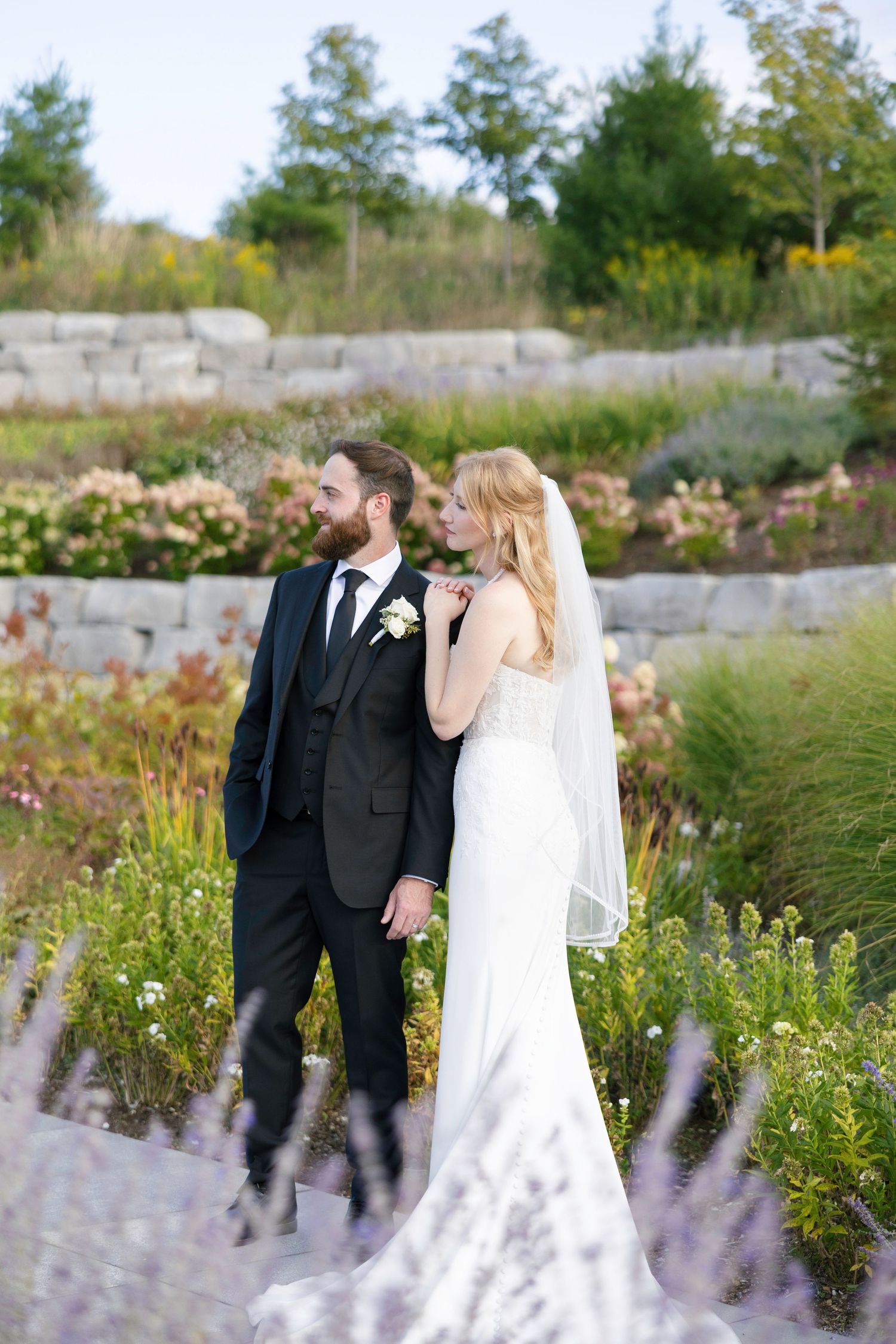 Bride and groom stand together in a lush garden, bride in white gown with veil, groom in dark suit with boutonniere.