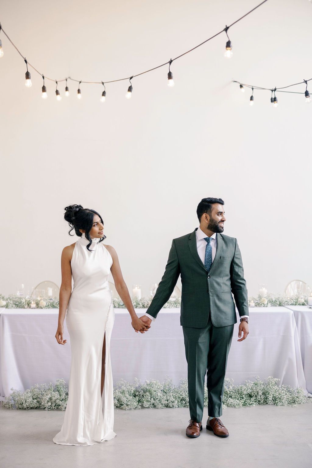 Elegant couple holds hands in modern venue, bride in sleek white gown, groom in green suit.