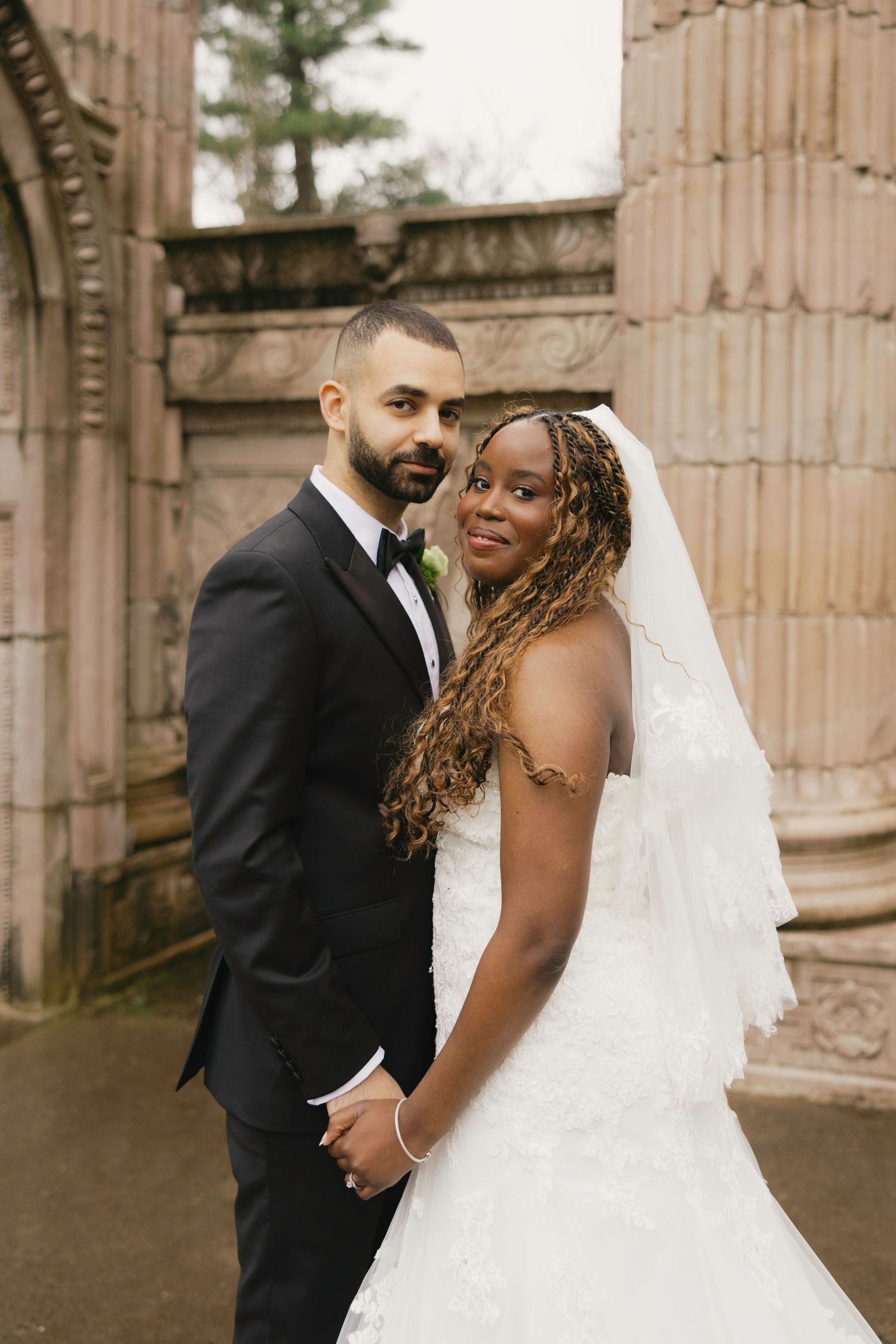 Interracial couple in wedding attire holding hands, bride in white lace gown with veil, groom in black tuxedo.