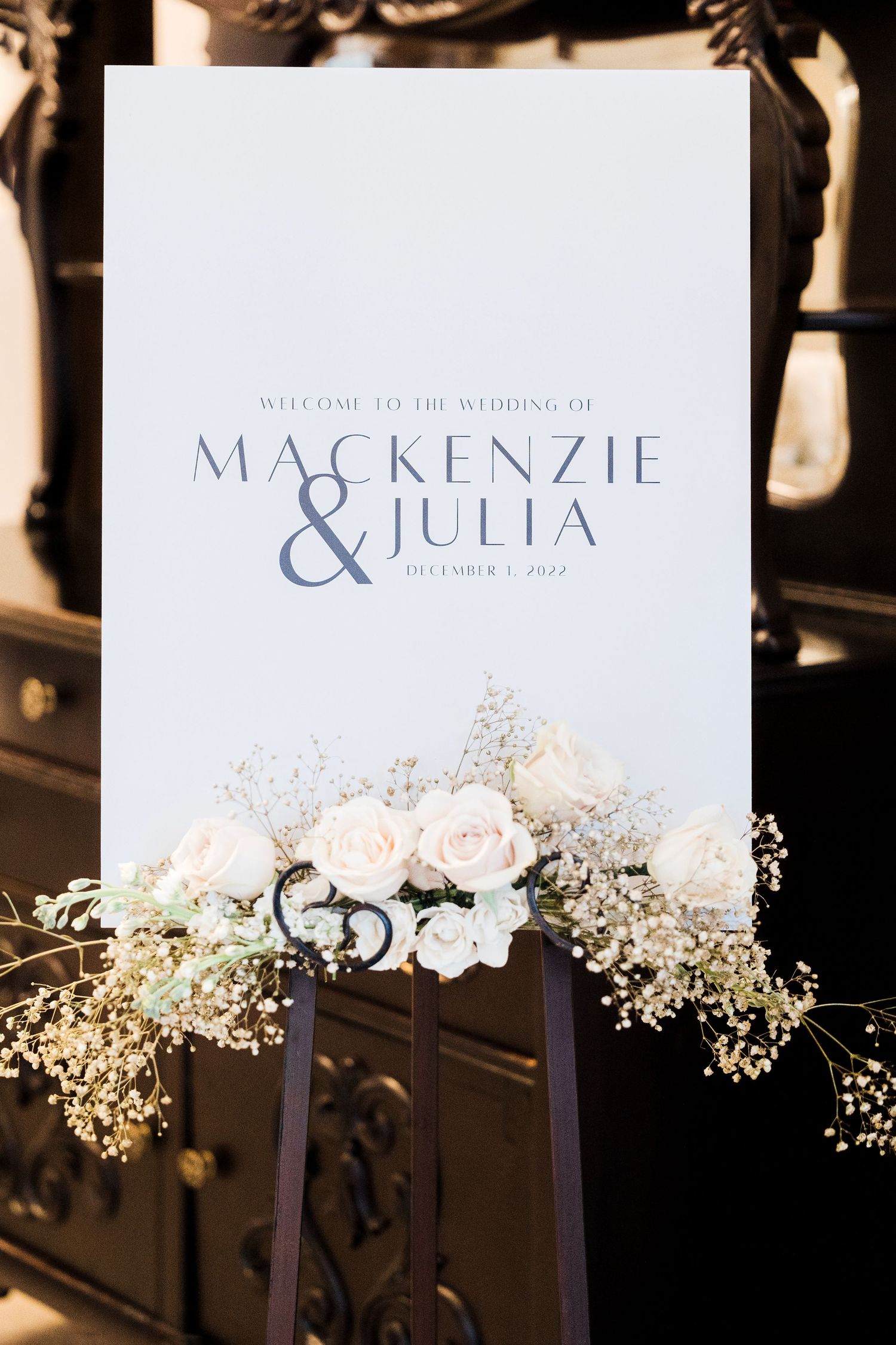 Elegant wedding welcome sign with white flowers and names Mackenzie & Julia displayed in modern typography.