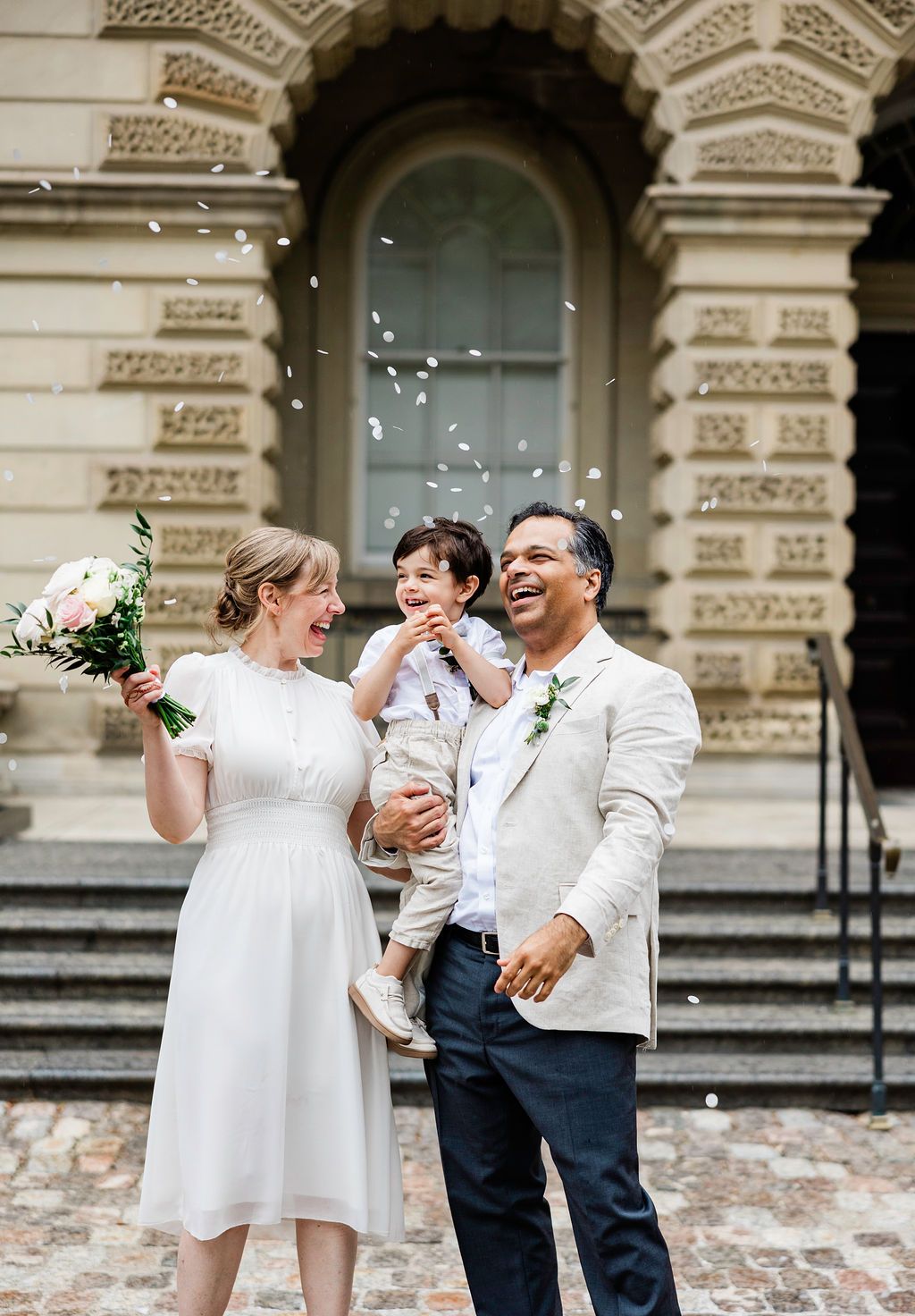 Happy couple with child celebrating wedding outside elegant building, bride holding bouquet with confetti falling.