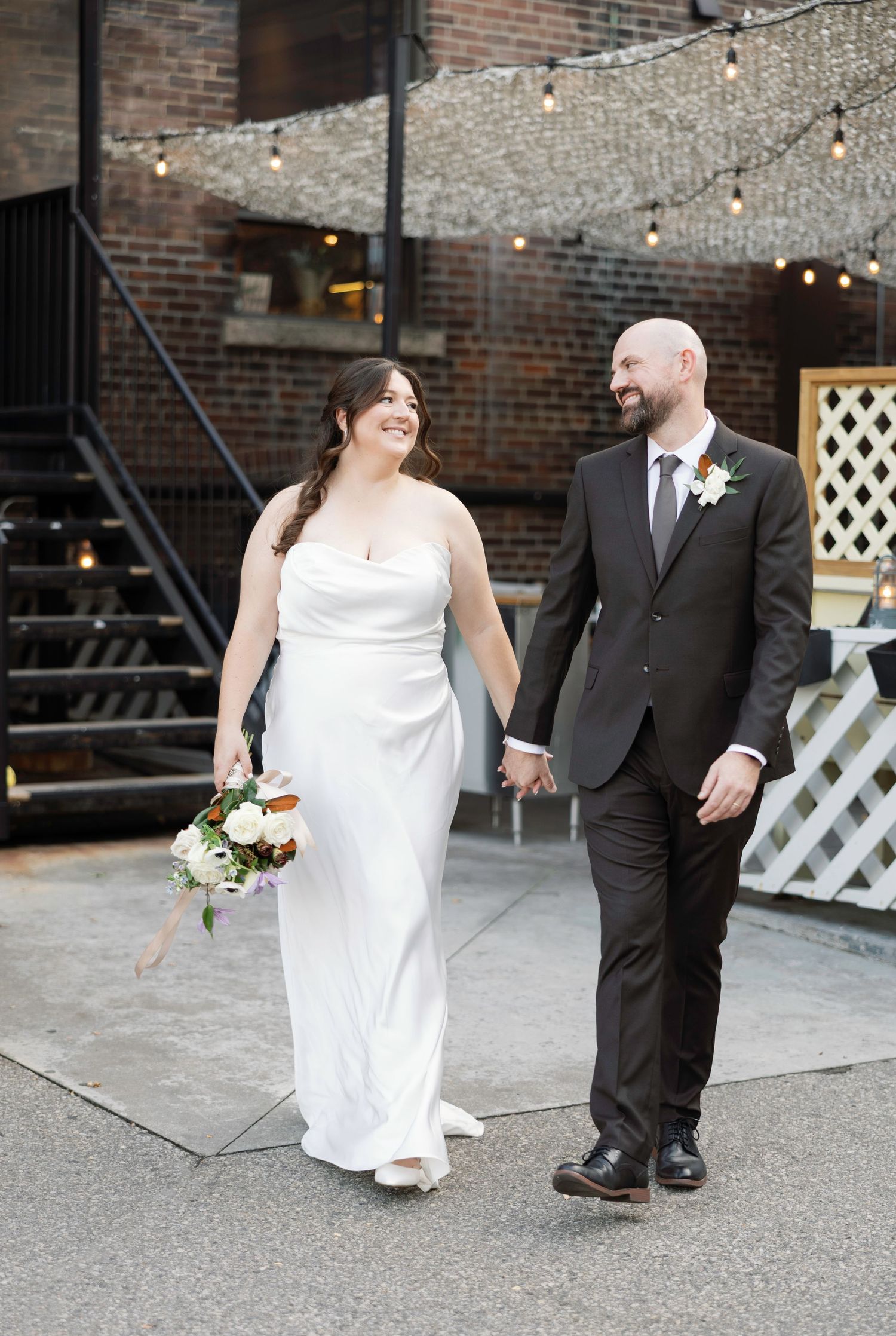 A bride in a white gown and groom in a dark suit hold hands and smile while walking in a brick courtyard.