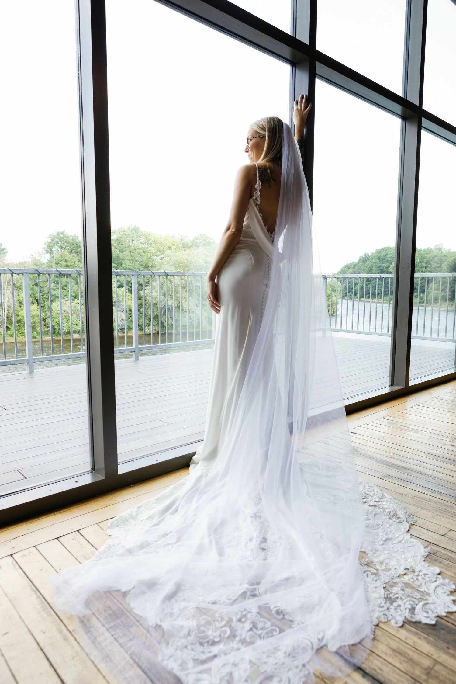 Elegant white wedding dress with flowing lace train displayed in a bright modern space with floor-to-ceiling windows.