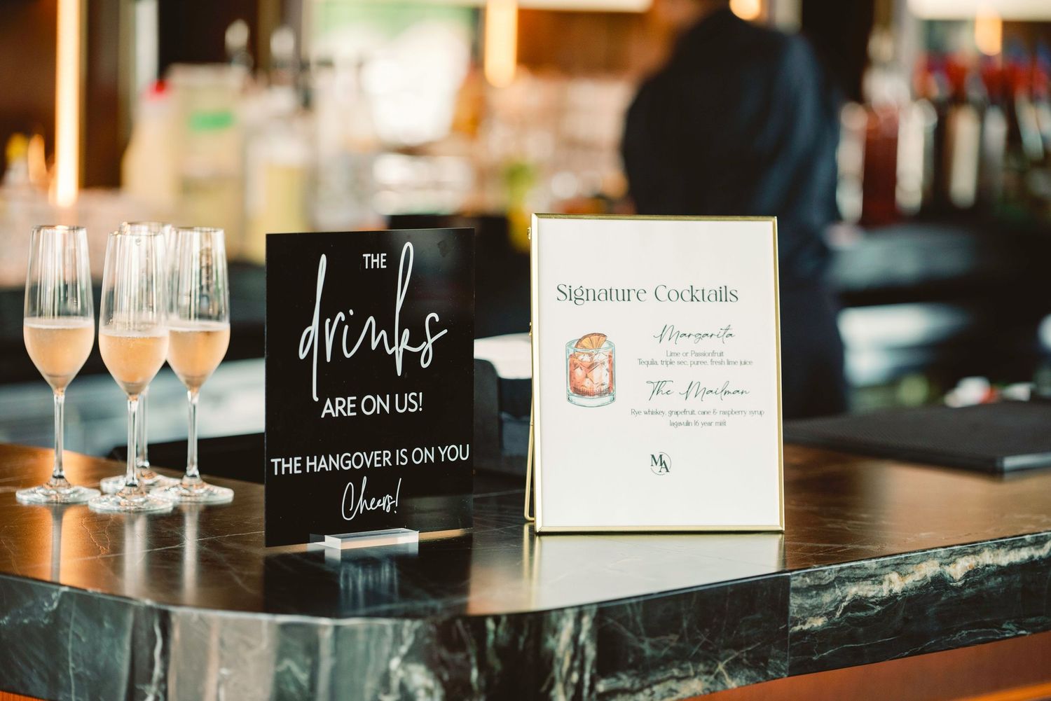 Wedding reception bar setup with drink menus and elegant glassware on dark wooden surface.