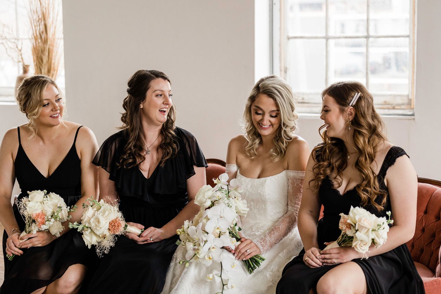 Bridesmaids in black dresses share joyful laughter while holding white floral bouquets in a bright indoor setting.