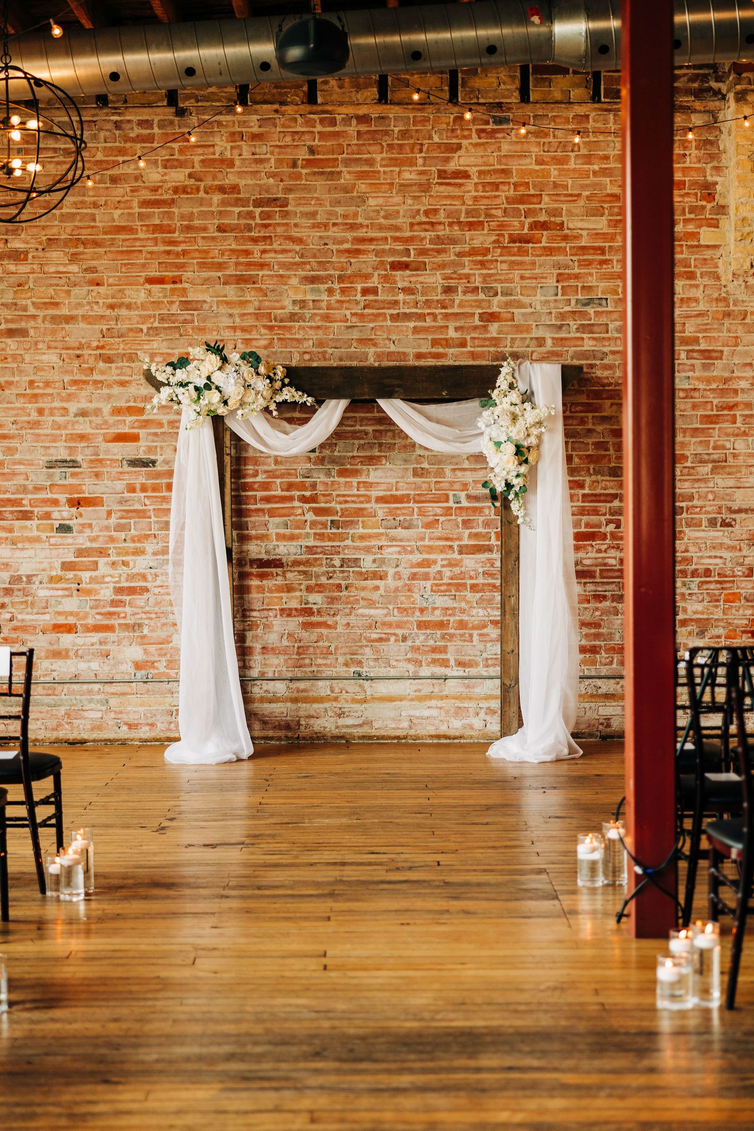 Elegant wedding ceremony arch with white fabric draping and floral arrangements against brick wall.