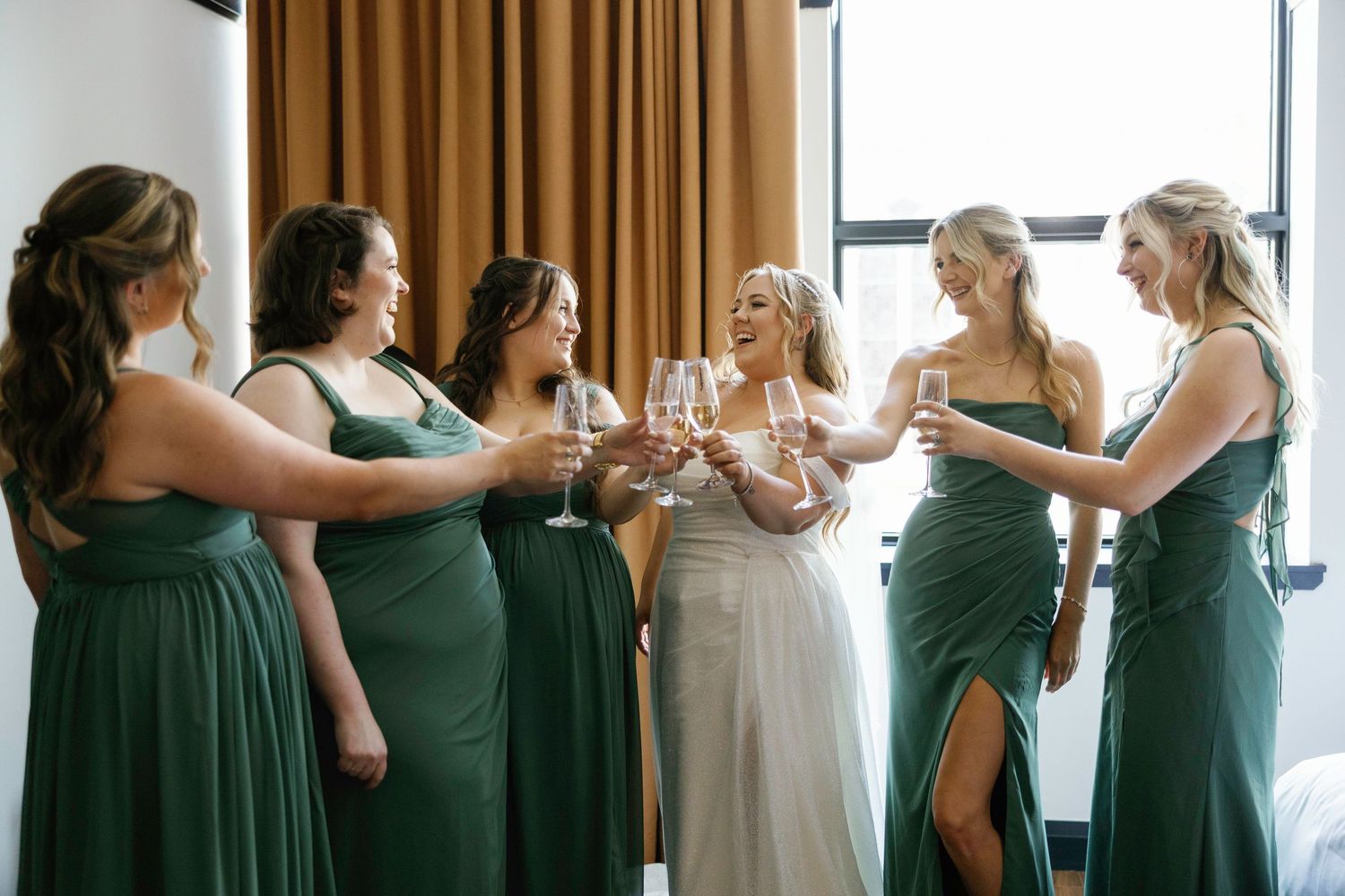 Bride in white gown toasting champagne with five bridesmaids in green dresses near a bright window.