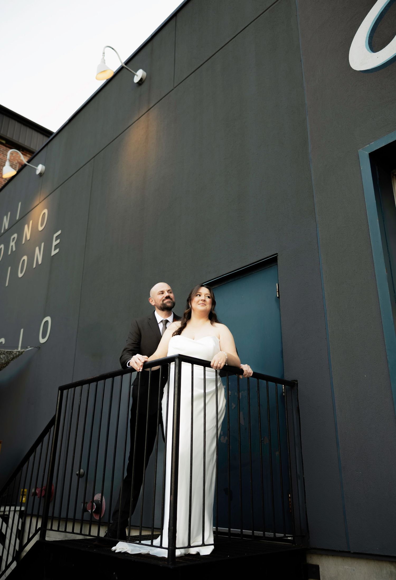Bride and groom stand on a balcony outside a dark modern building, gazing upward in wedding attire.