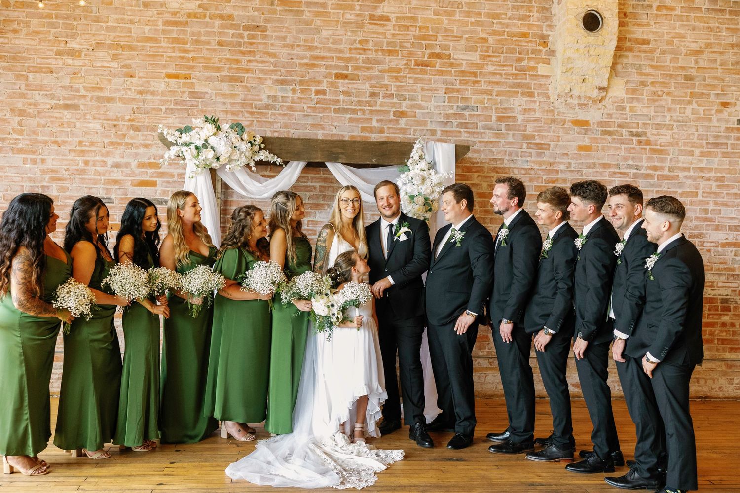 Wedding party members in formal attire lined up against rustic brick wall with elegant floral arch decoration.