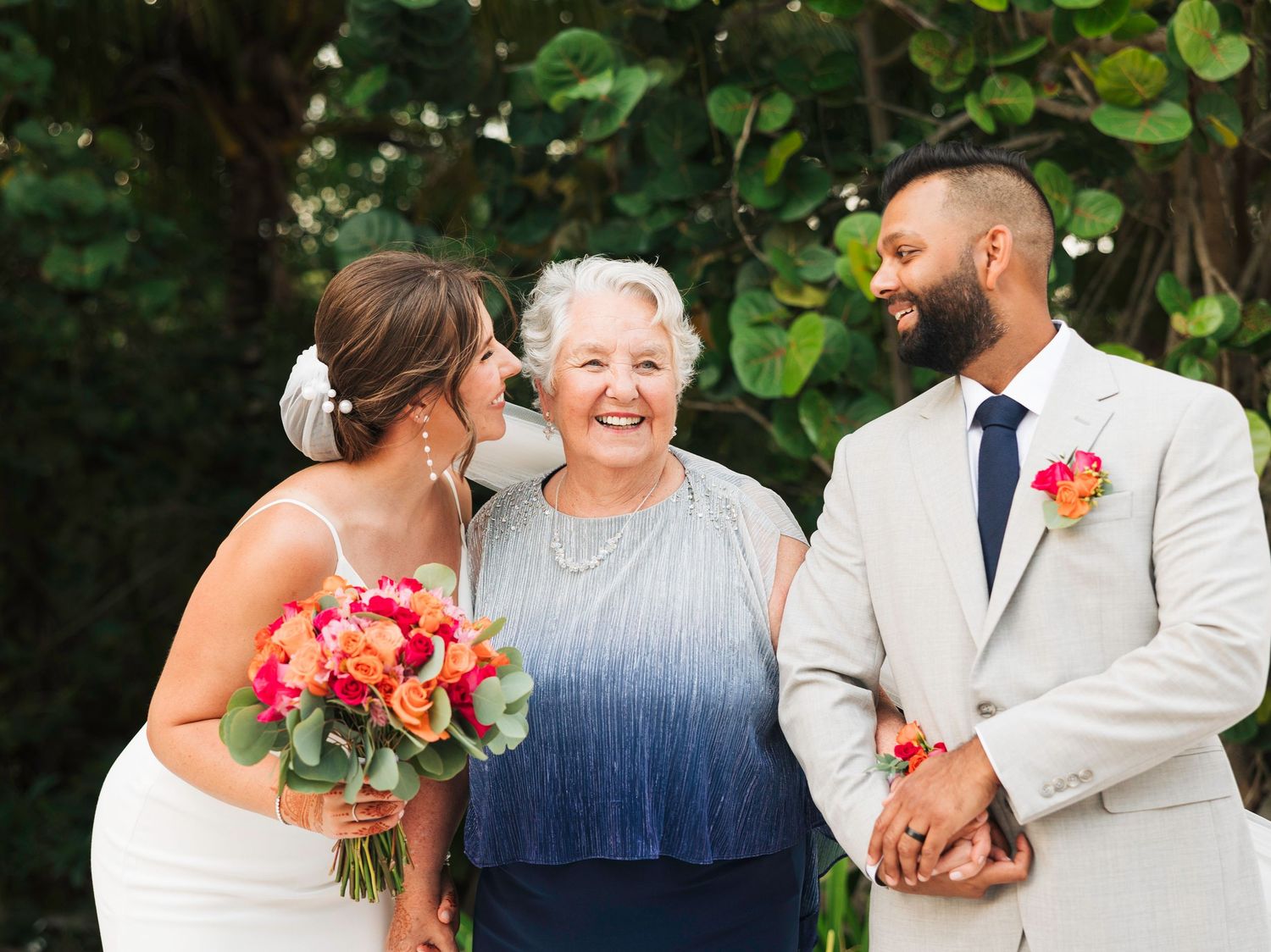 Wedding party poses outdoors with vibrant coral bouquets against lush green foliage backdrop.