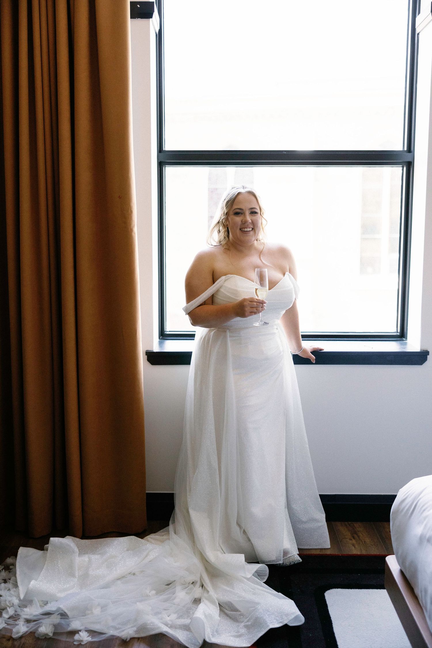 A bride in a flowing white wedding gown stands by a window with natural light streaming in.