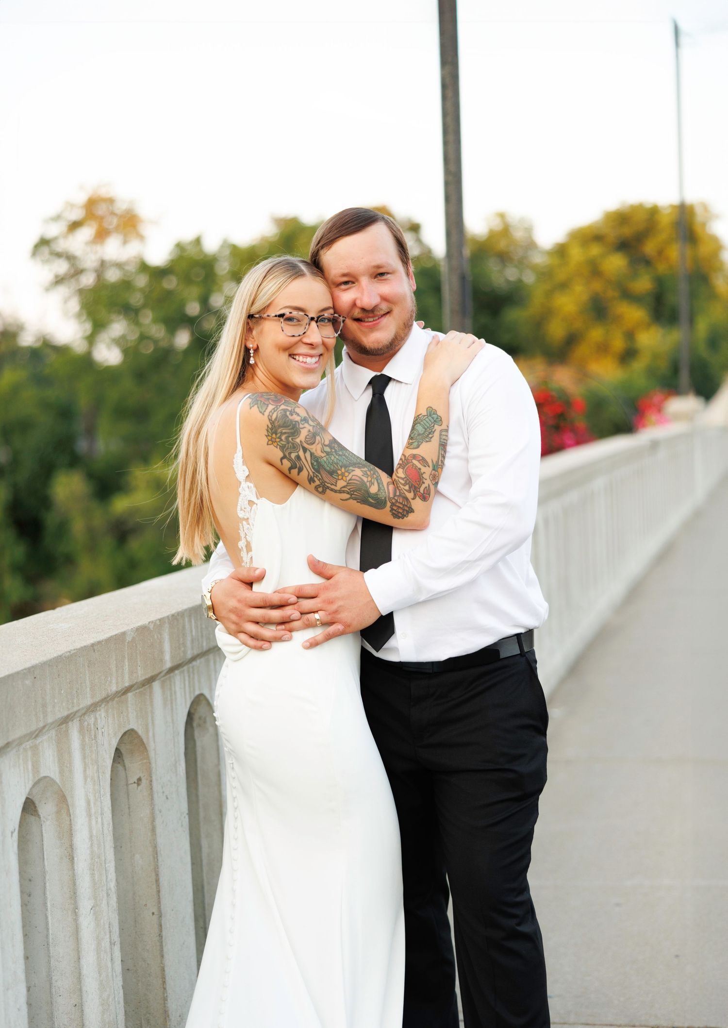 A couple in wedding attire embraces on a white bridge during an autumn day with fall foliage in the background.