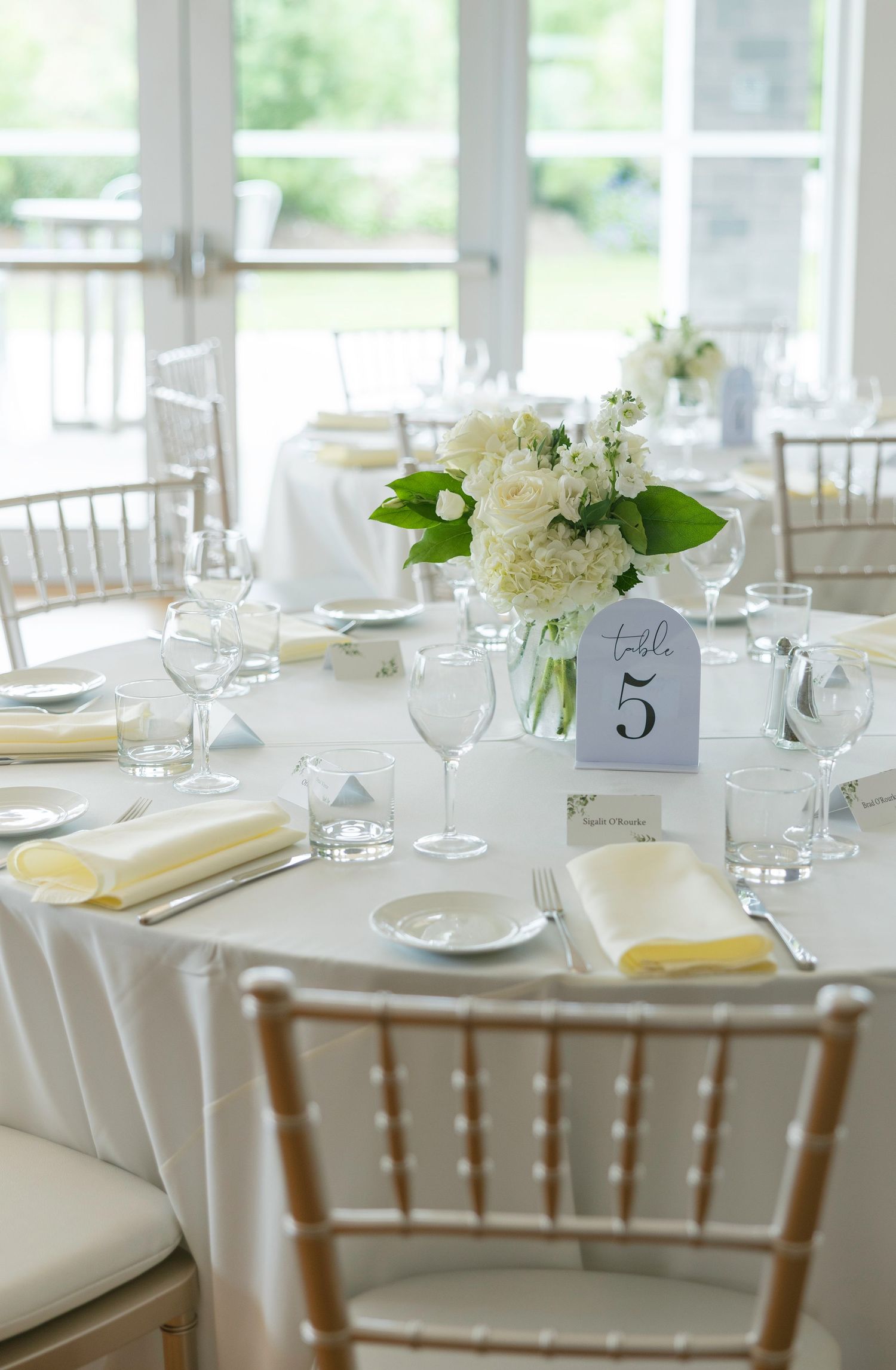 Elegant wedding reception table setting with white linens, chiavari chairs, floral centerpieces and table number.