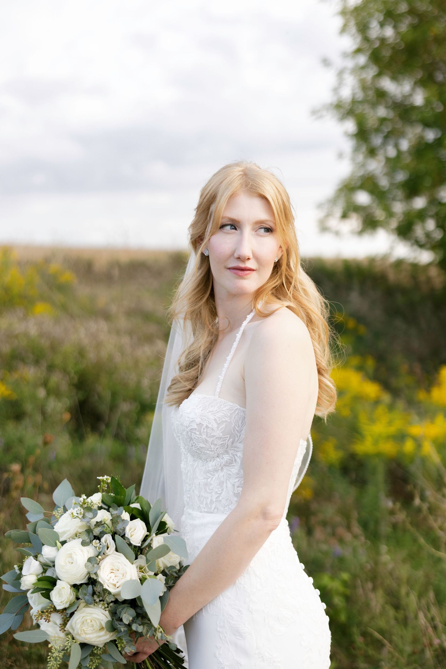 A bride in a white lace dress holds a bouquet of white flowers and greenery in an outdoor garden setting.
