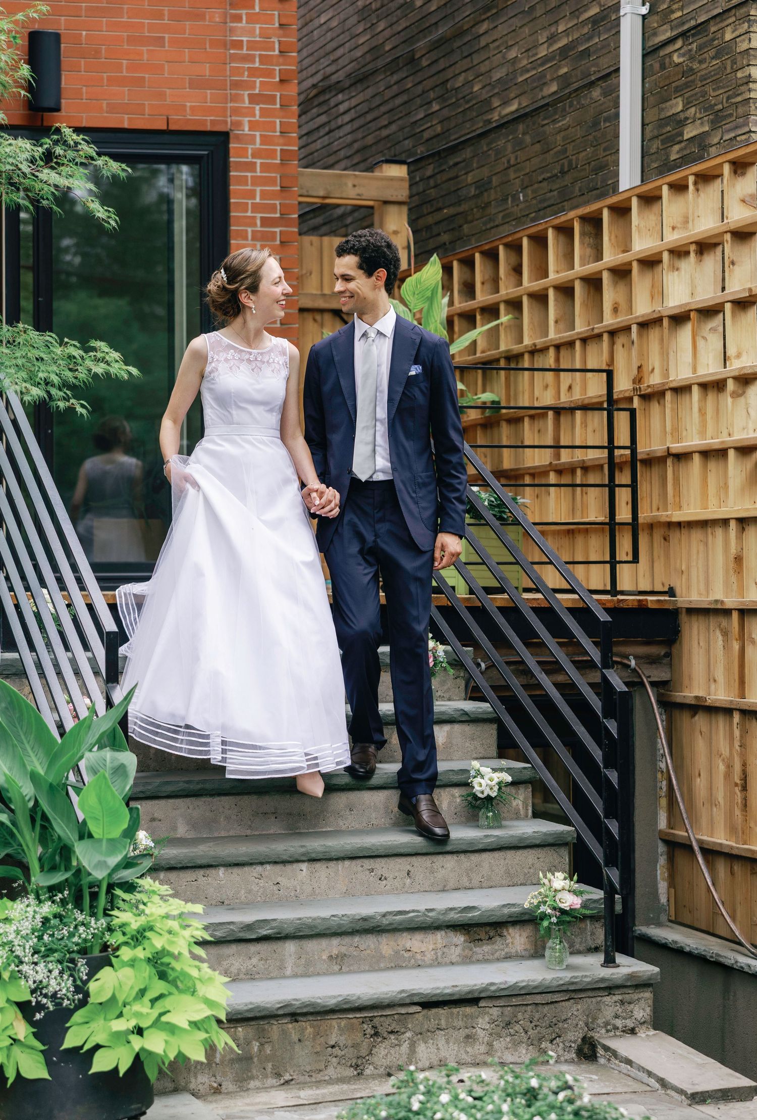 Newlyweds posing on concrete steps with wooden fence and brick wall backdrop.