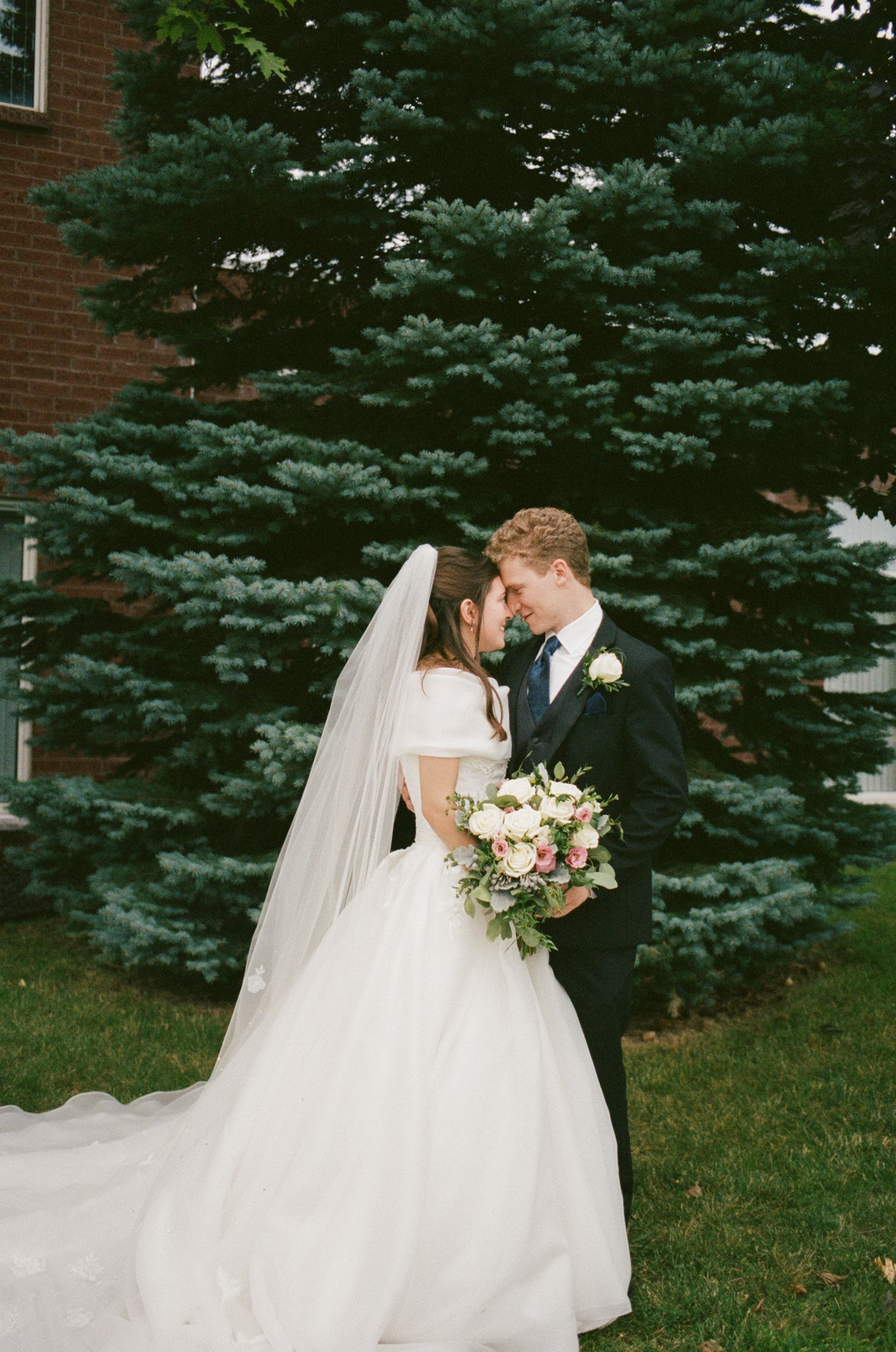 A bride and groom share a romantic moment in front of a lush evergreen tree, bride holding a bouquet in a white gown.