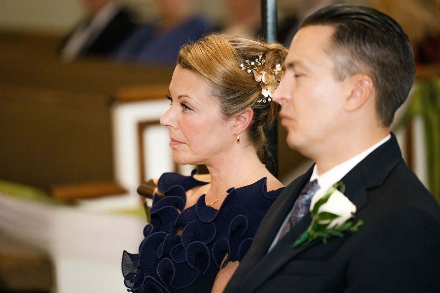 A woman with an updo and a man in a tuxedo sit together during a wedding ceremony in a church.