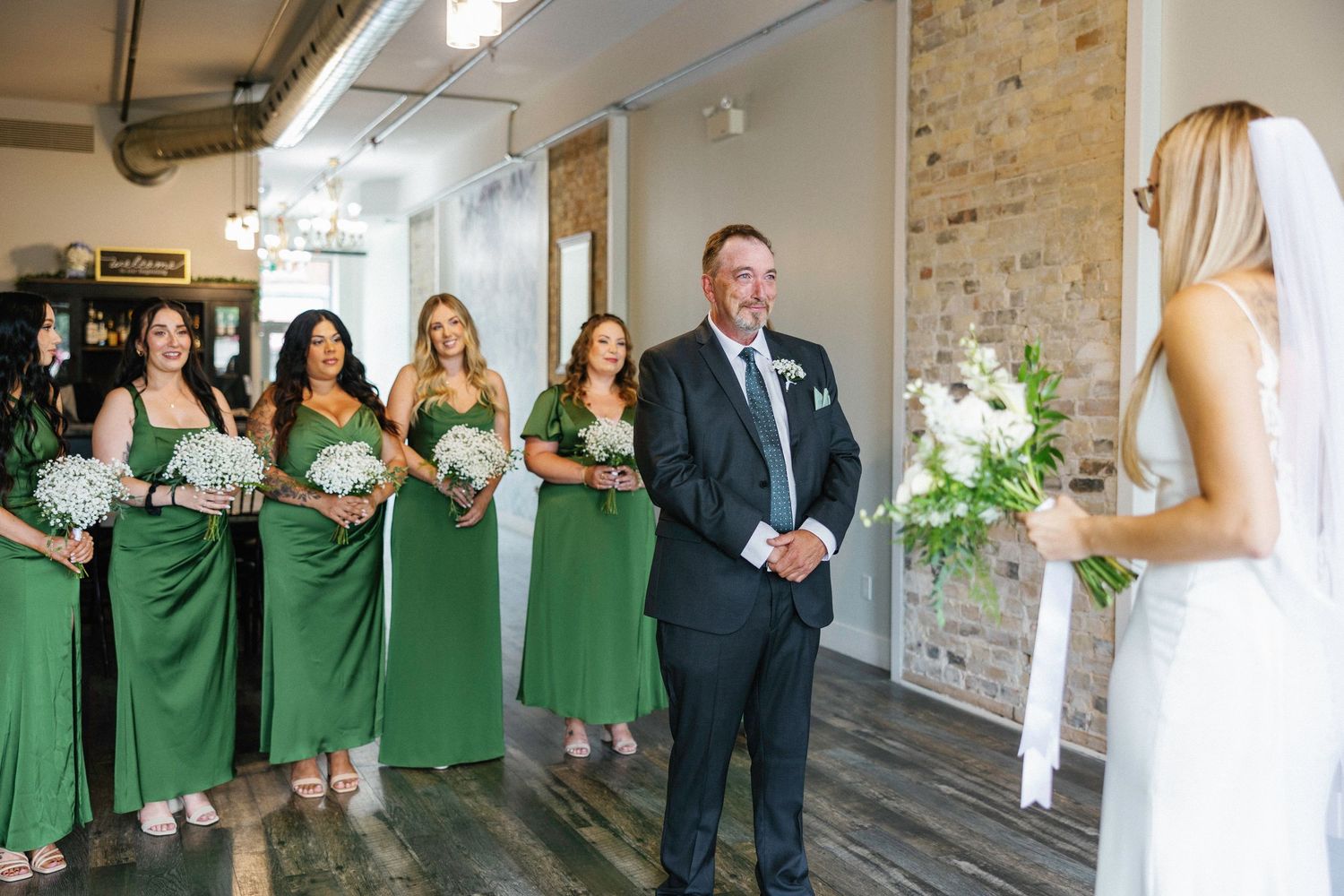 Bridesmaids in emerald green dresses and bride in white gown stand together during wedding ceremony at indoor venue.