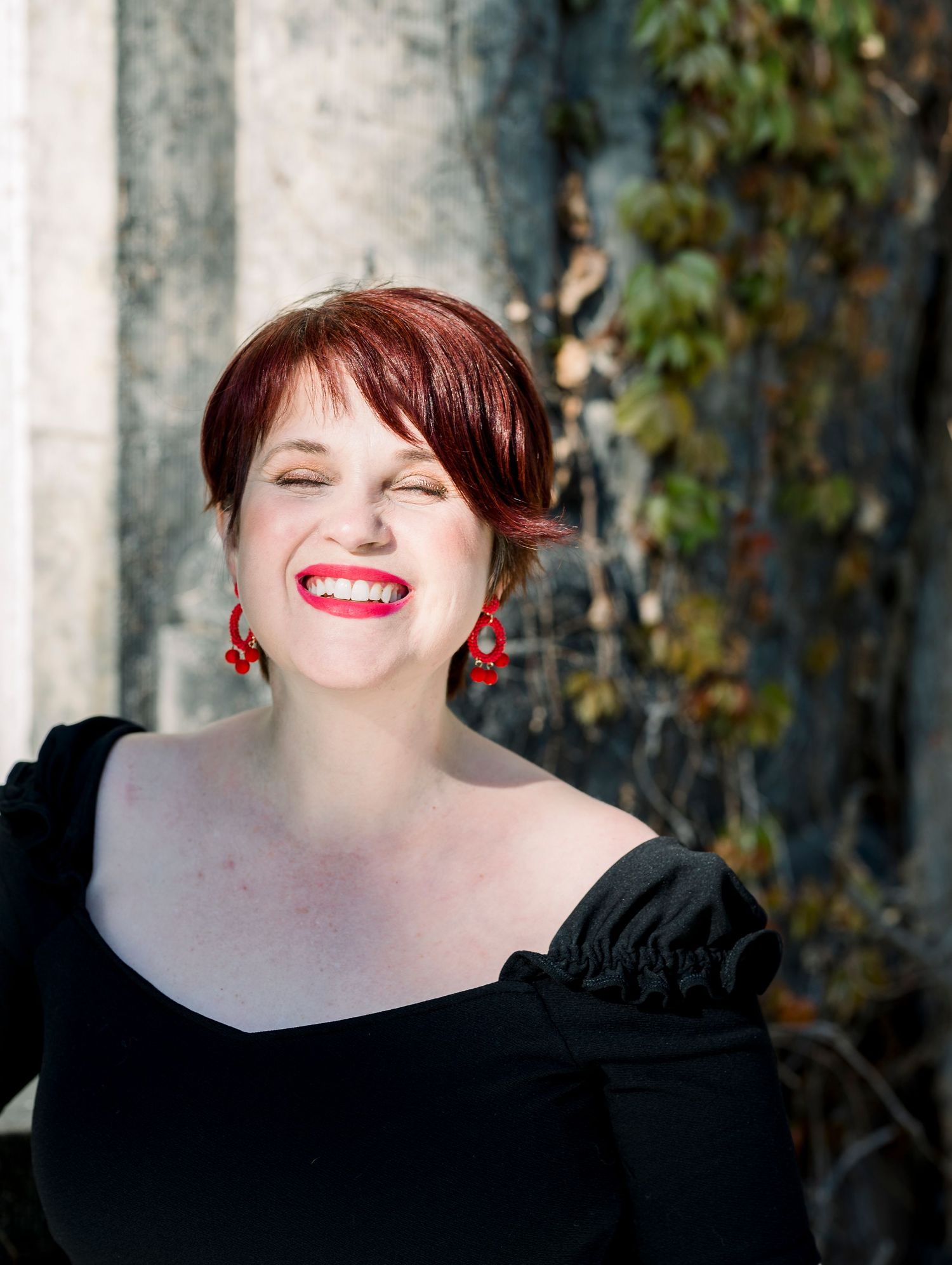 A laughing woman with short red hair and red earrings wearing a black off-shoulder top, posing outdoors near a stone wall.