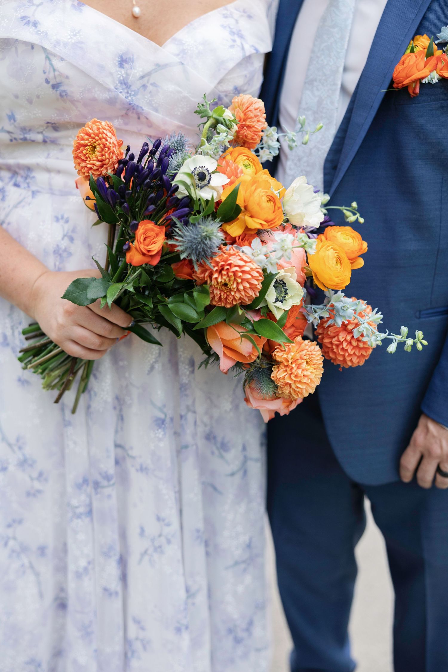 Bride in floral dress holding vibrant orange and blue wedding bouquet with dahlias, ranunculus, and anemones beside groom in navy suit.