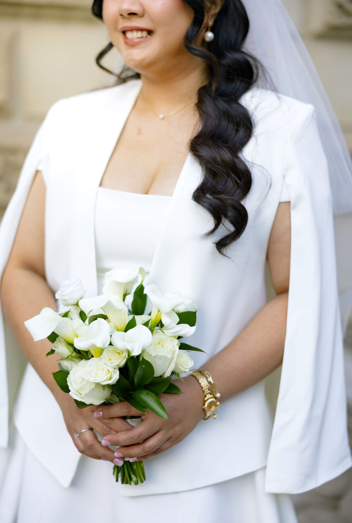Bride in white wedding dress with cape sleeves holds elegant bouquet of white roses and calla lilies, wearing gold bracelet.