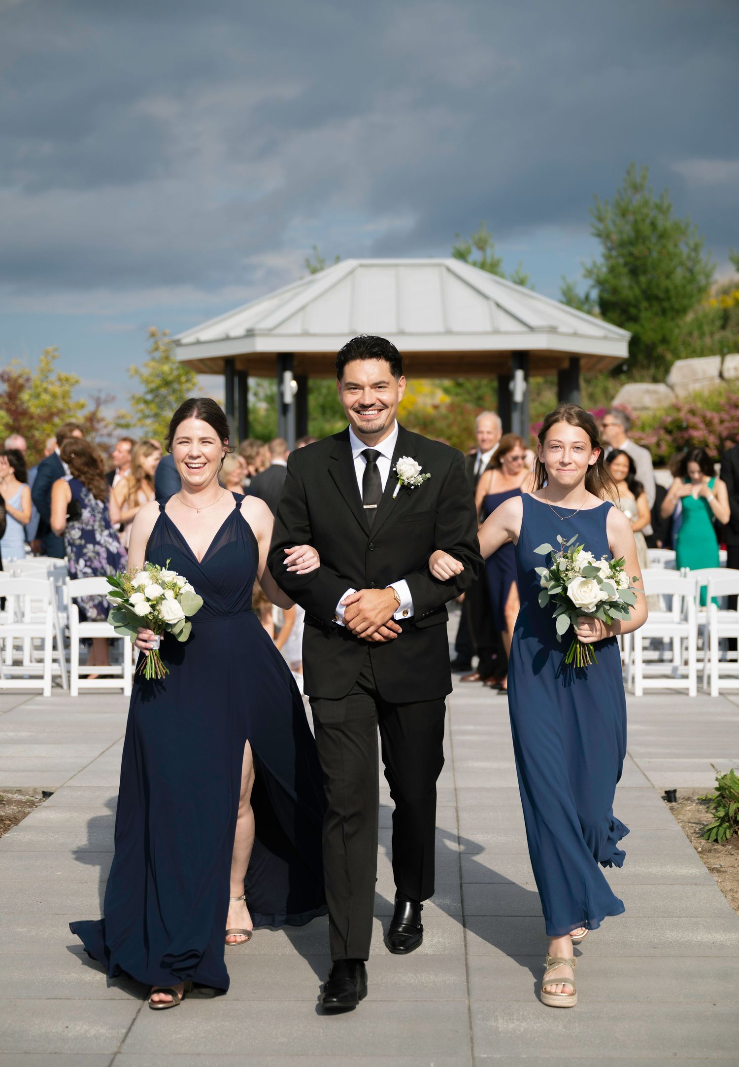 Wedding party members in navy blue dresses and dark suits walk and celebrate together on an outdoor patio after ceremony.
