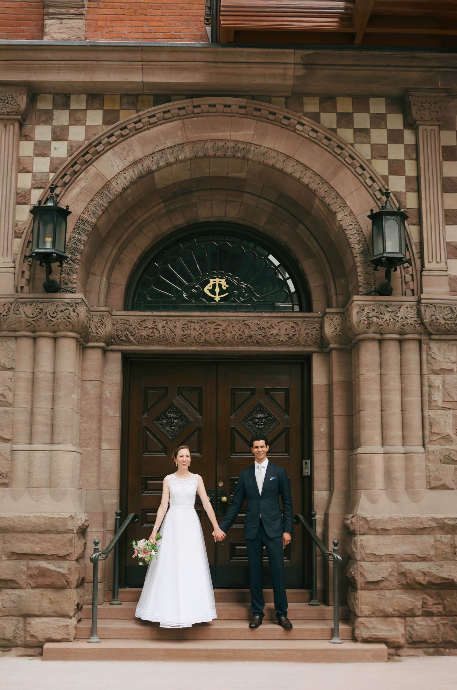 A couple in wedding attire pose in front of an ornate stone archway entrance with decorative columns.