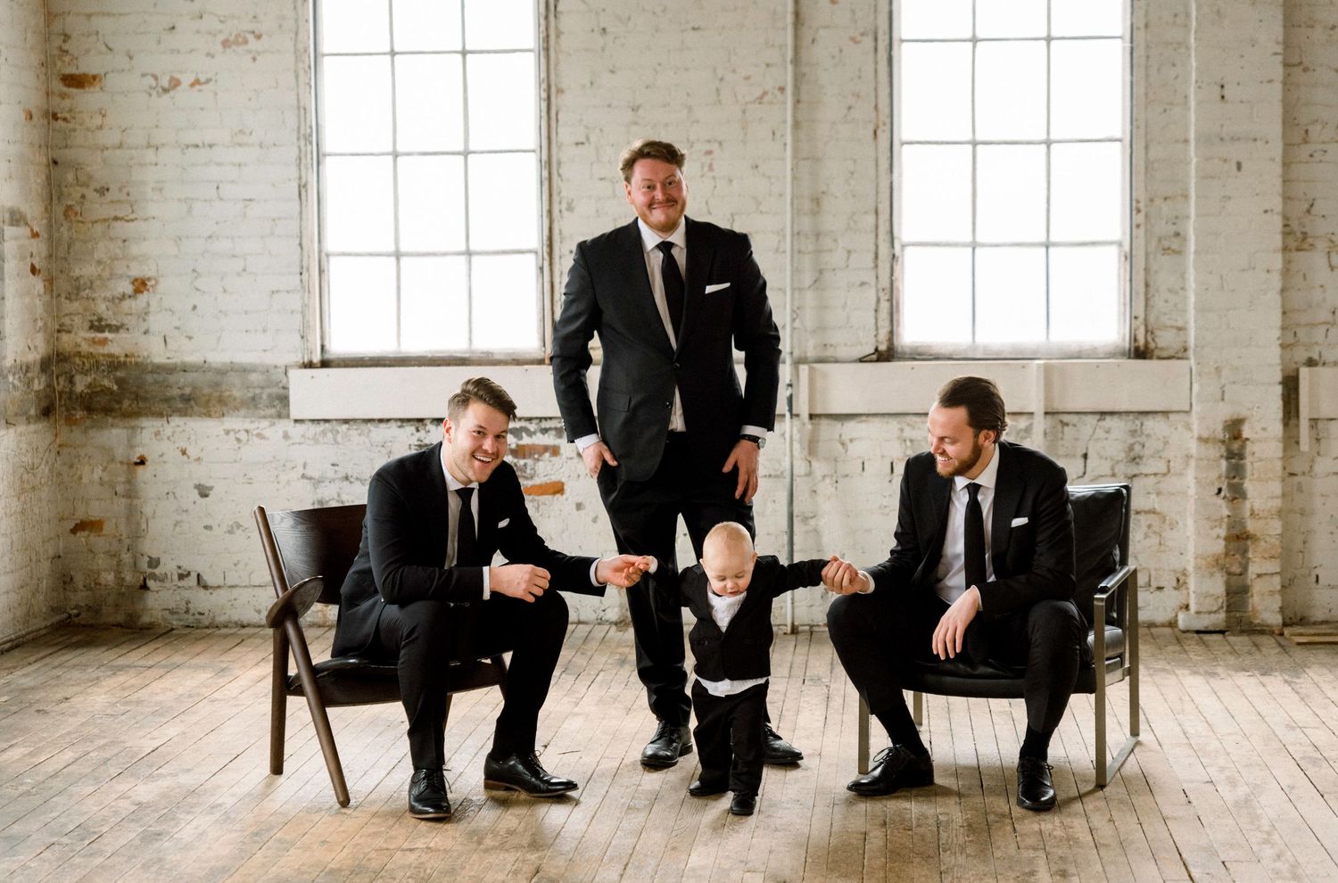 Wedding party members in dark suits pose playfully in front of bright windows in rustic venue.