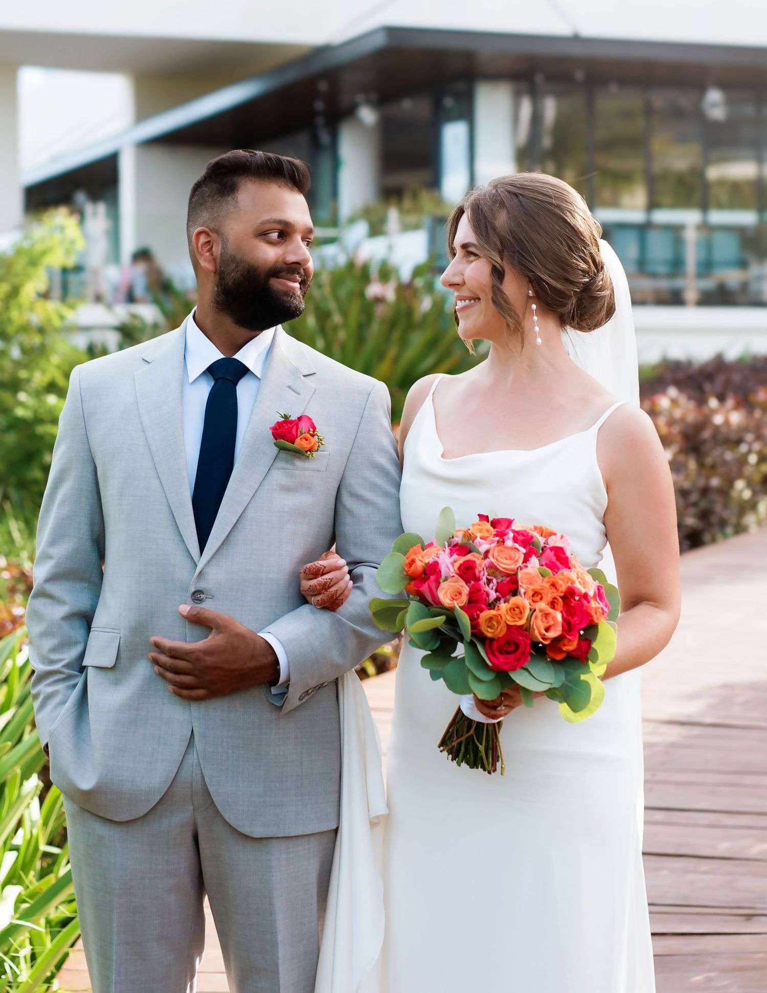 Elegant wedding couple in light gray suit and white dress holding coral and pink bouquet outdoors.