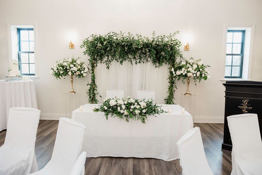 Modern white wedding ceremony space with natural light from windows and greenery decorations.