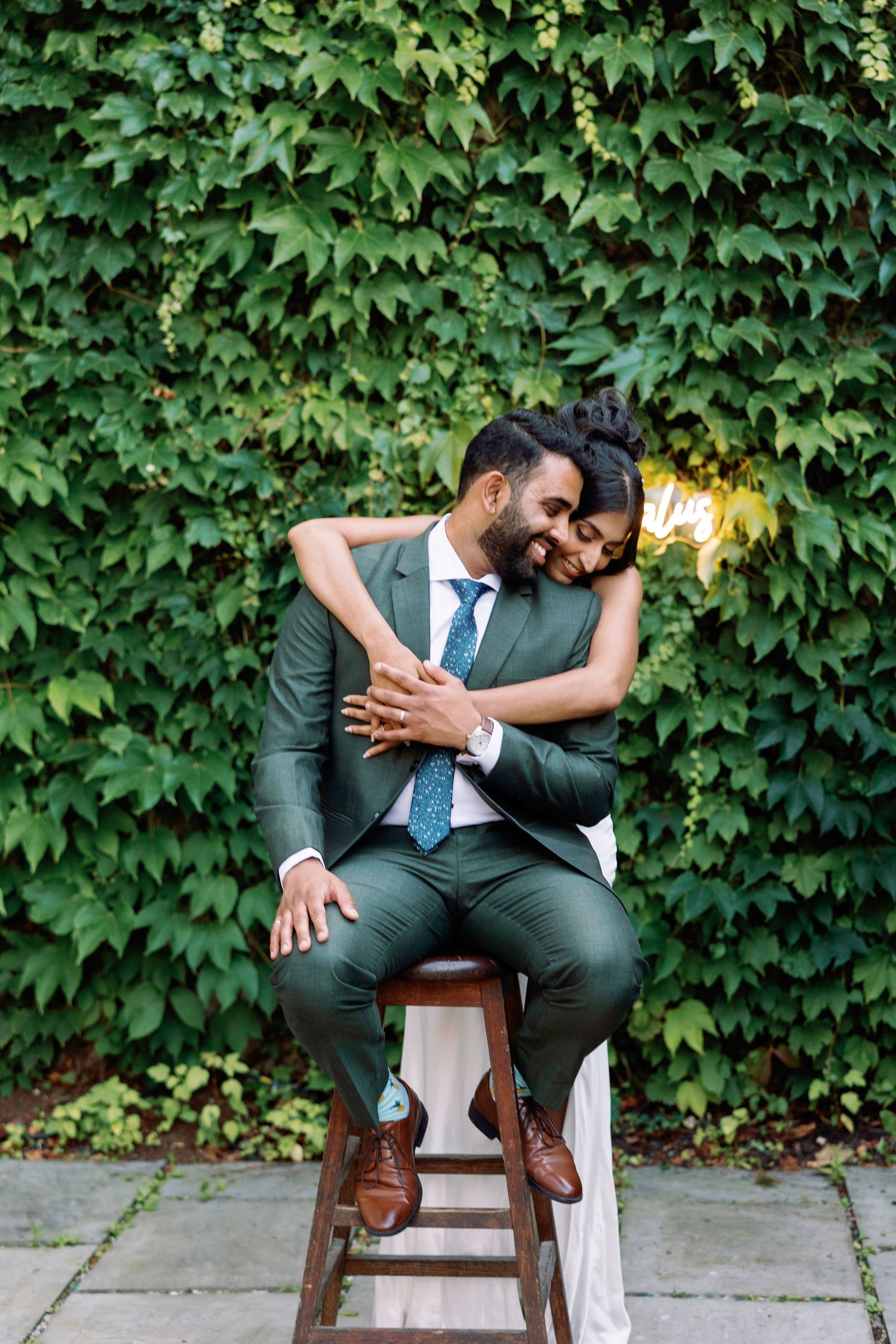 Wedding couple shares playful moments while seated on wooden chairs against an ivy wall.