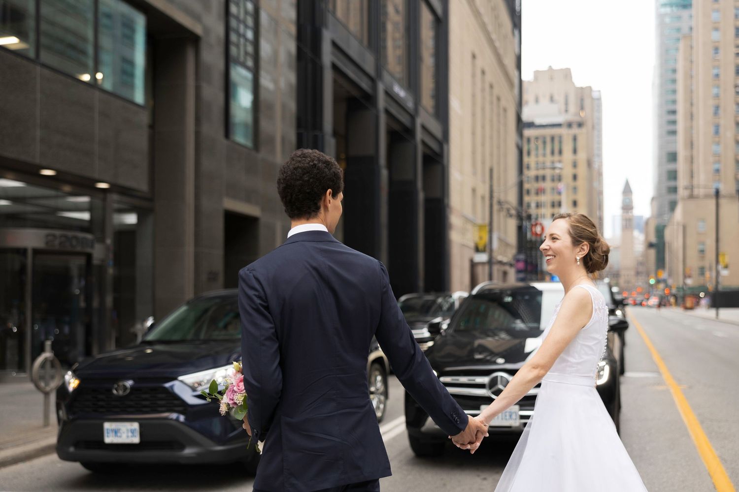 A couple in wedding attire walks through an urban street with tall buildings in the background.