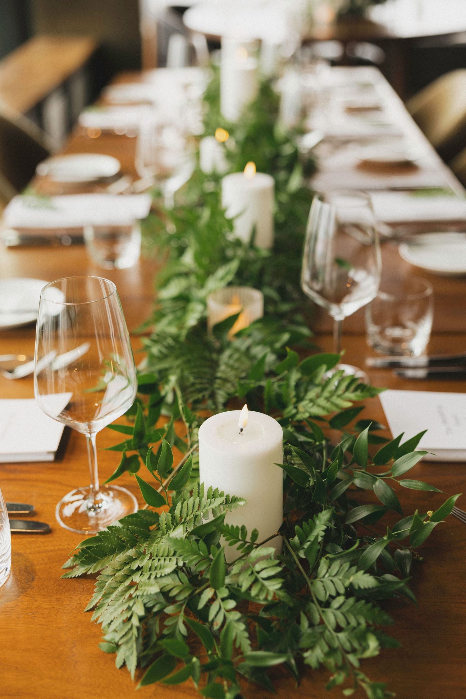 Elegant table setting with greenery garland, white candles, and wine glasses on wooden surface.