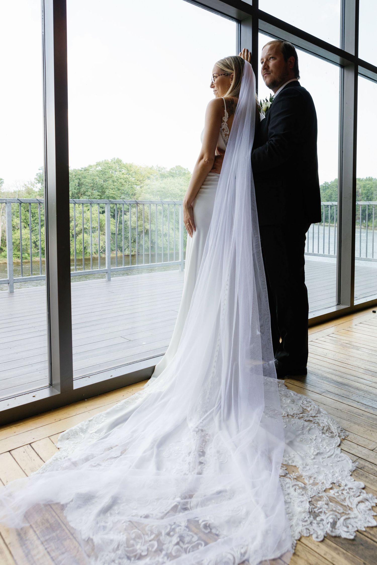Elegant wedding couple stands by large windows with flowing white cathedral veil trailing behind on hardwood floor.