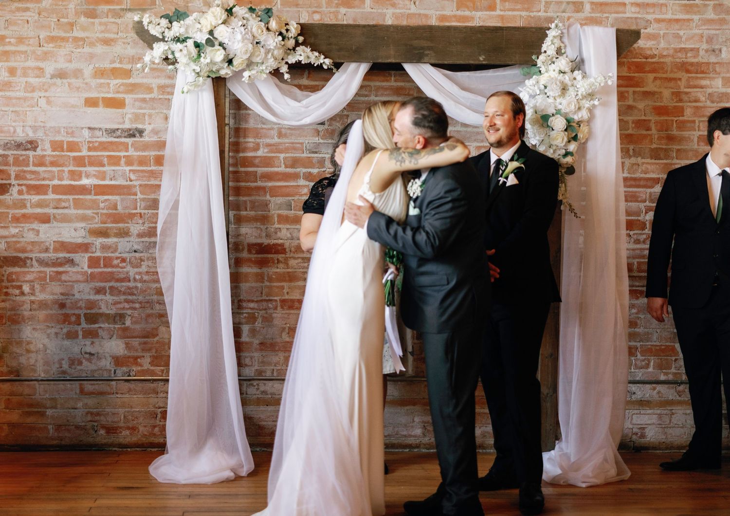 Wedding ceremony taking place under white draped arch with floral decorations in rustic brick venue.