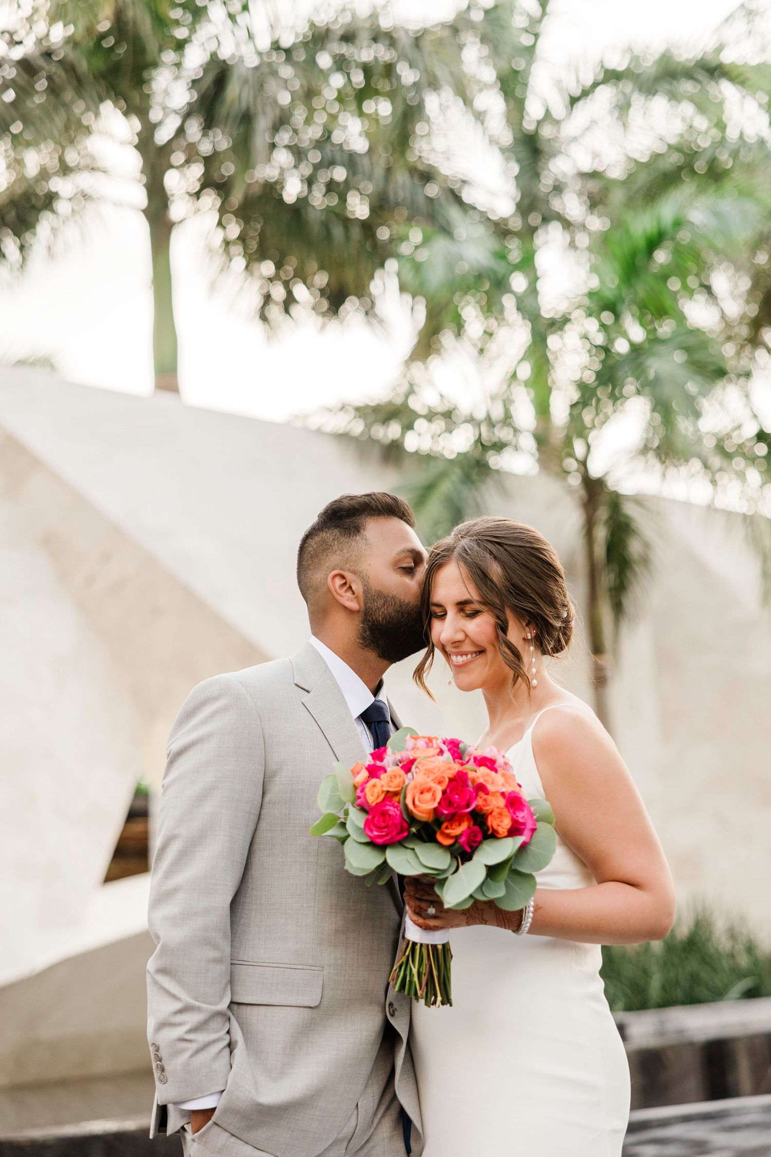 Groom in gray suit kisses bride's cheek as she smiles holding vibrant pink and orange bouquet outdoors.