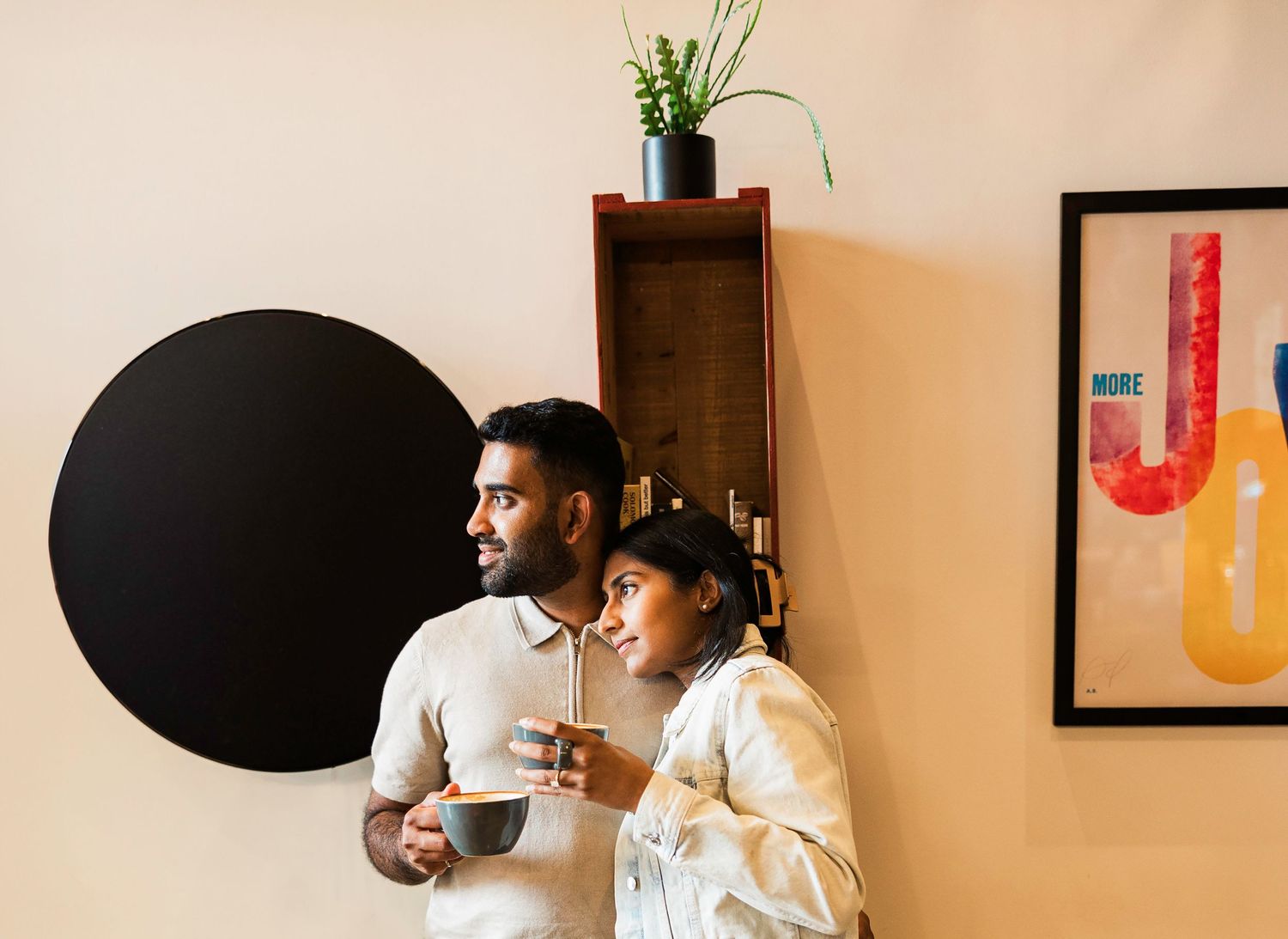 A couple stands together holding coffee cups, gazing thoughtfully in a modern room with minimalist decor.