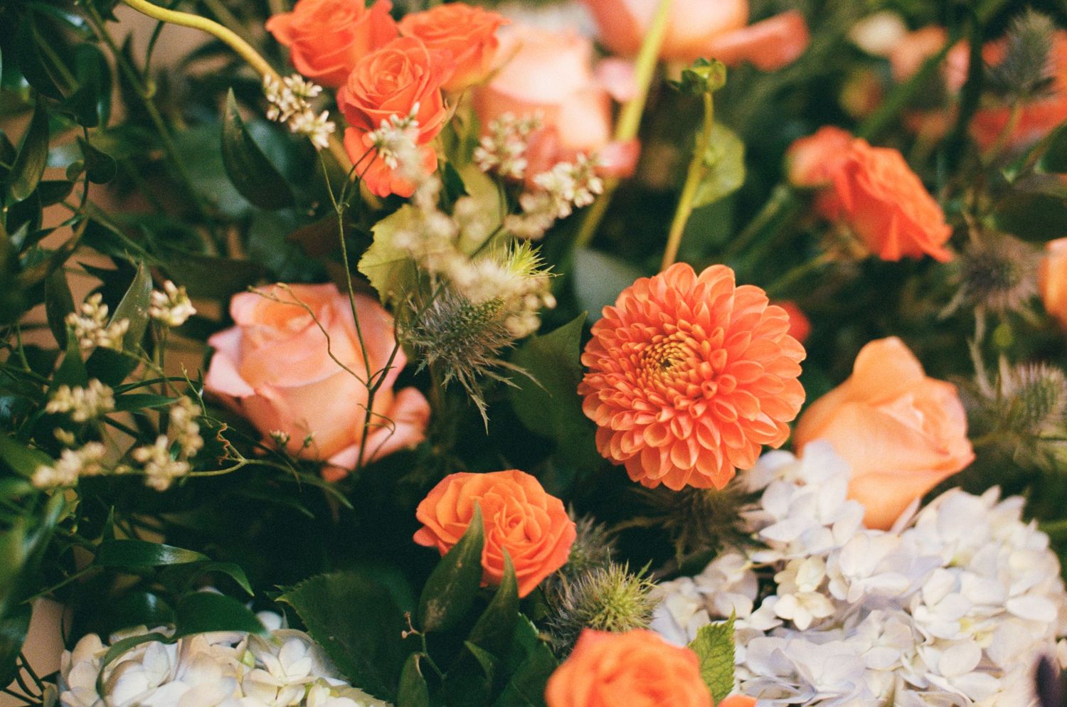 Vibrant orange dahlias and roses arranged with white hydrangeas and lush greenery in an elegant floral bouquet.
