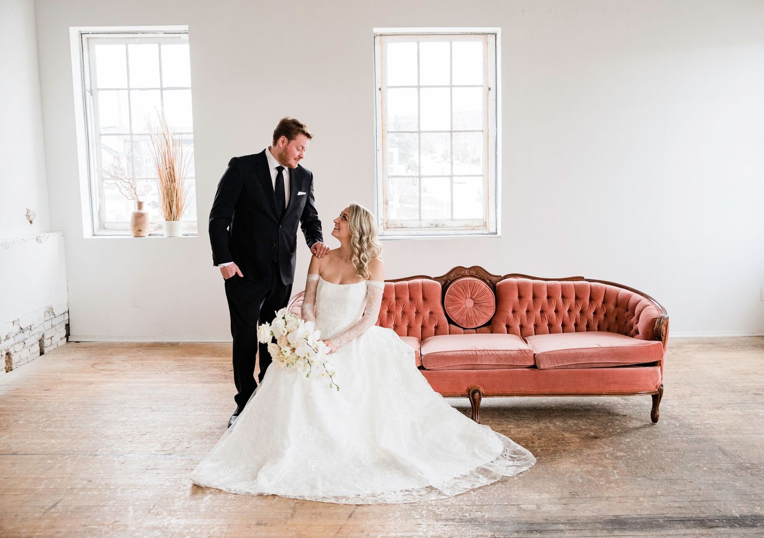 Elegant wedding couple poses in a bright white room with vintage pink couch and large windows.