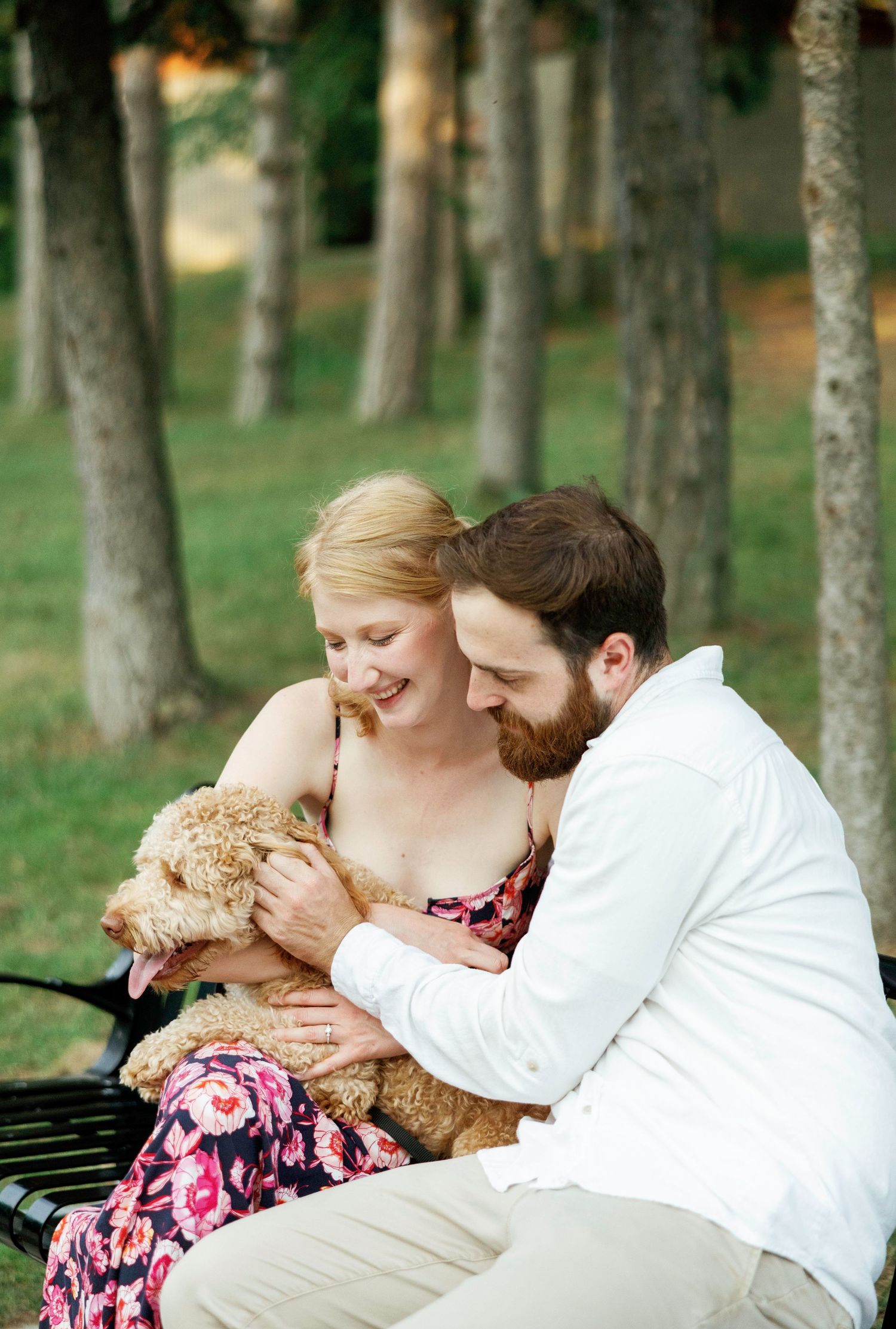 Couple sitting on bench with goldendoodle puppy in wooded setting.