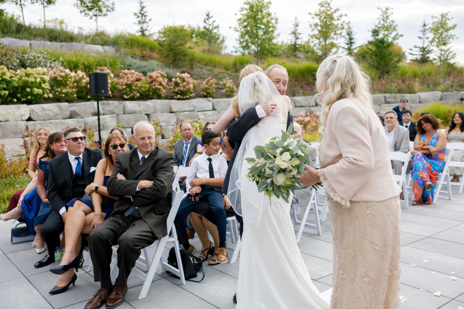 Outdoor wedding ceremony on stone patio with guests seated on white chairs and floral garden backdrop.