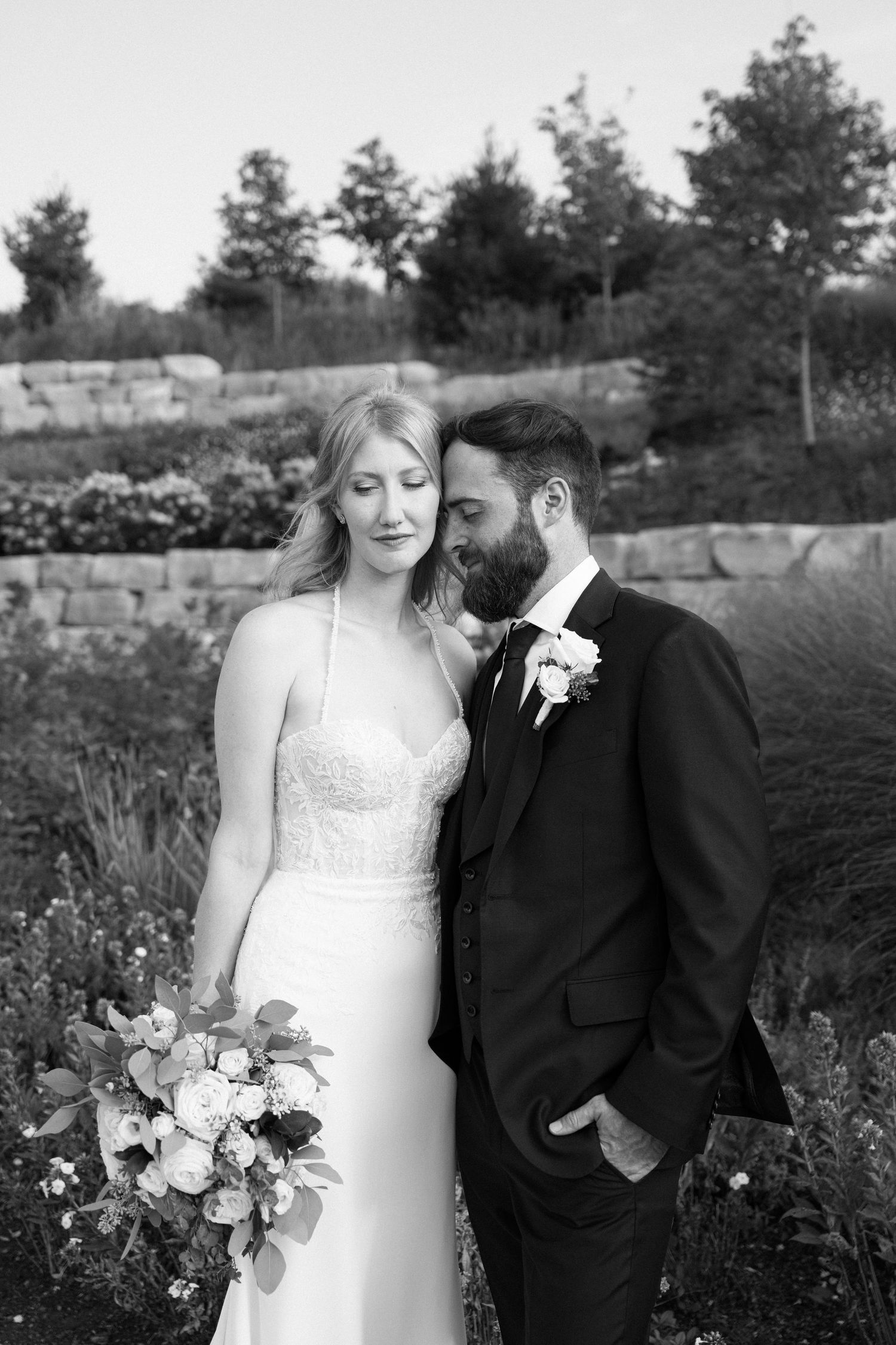 Black and white wedding portrait of a couple embracing in a garden setting with stone walls and wildflowers.