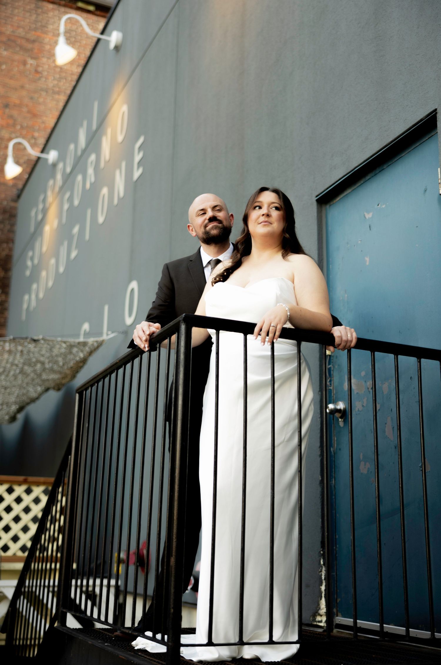 Bride in white gown and groom in dark suit pose on urban balcony with brick wall backdrop.