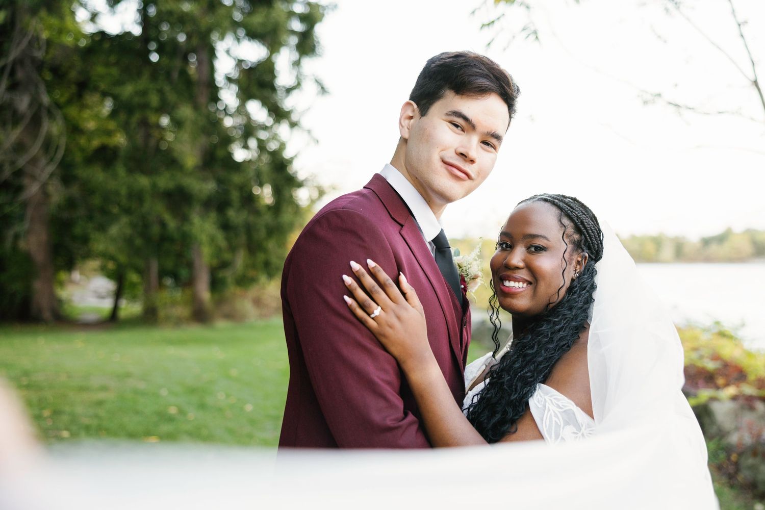 Interracial couple posing outdoors on wedding day, groom in burgundy suit, bride in white veil and lace dress.