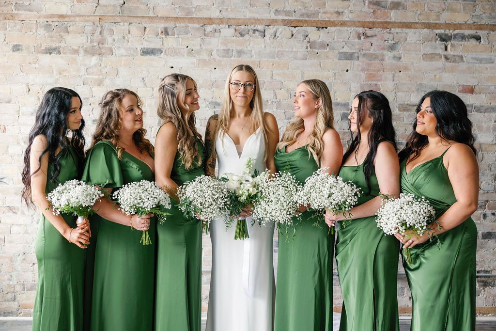 Bride in white gown with six bridesmaids in green dresses holding white baby's breath bouquets against brick wall.