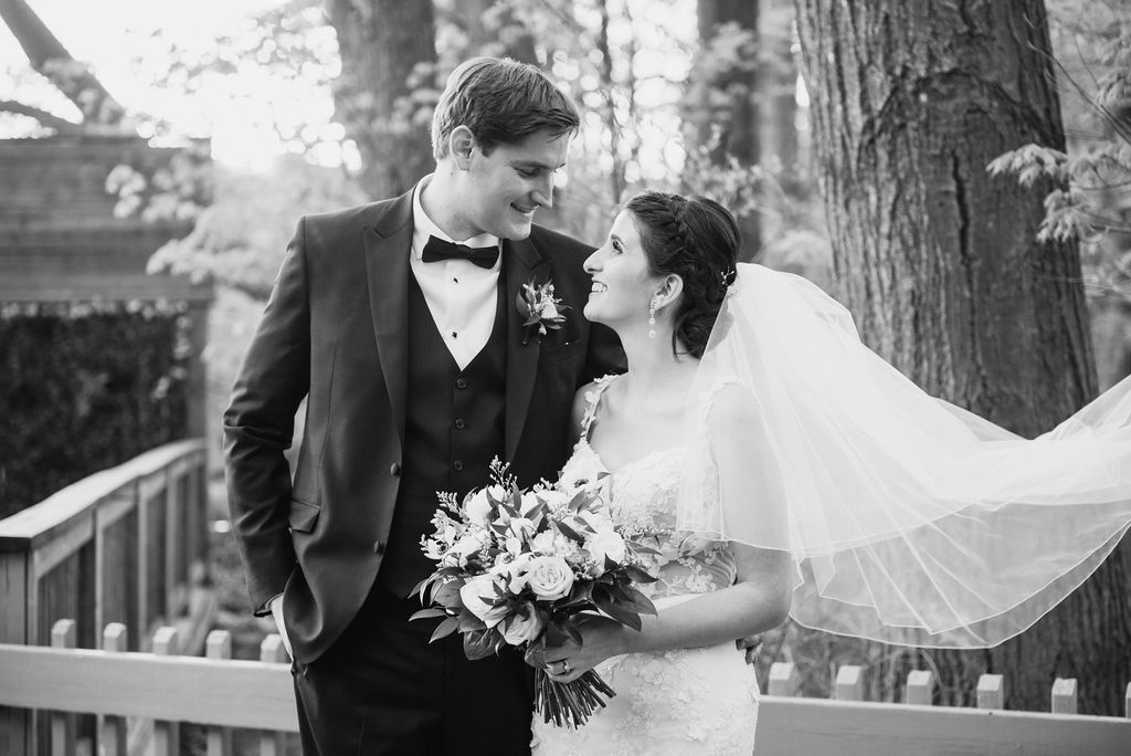 A bride and groom walk hand in hand through a scenic outdoor garden path surrounded by trees.