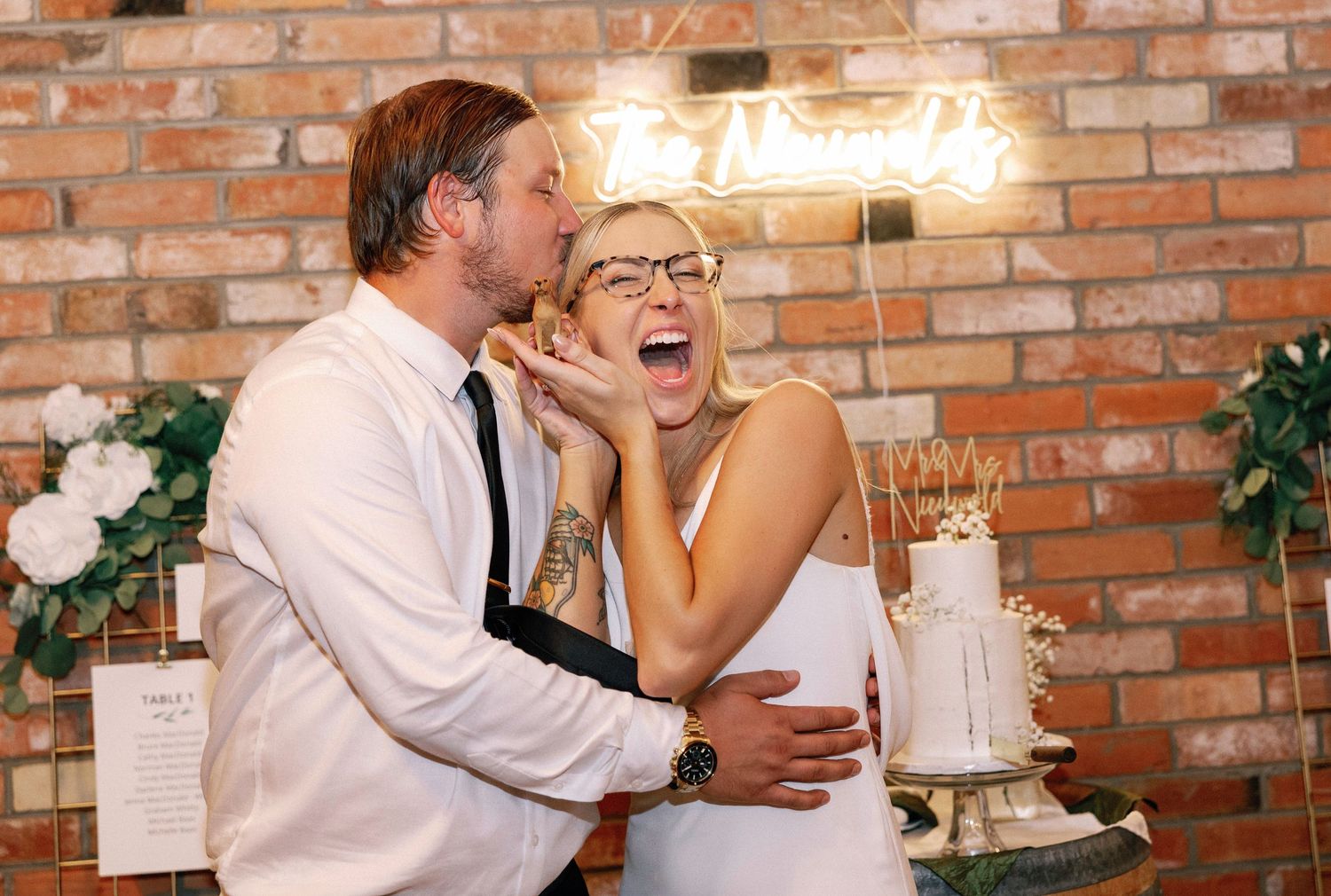Joyful couple shares an intimate moment during their wedding reception against a rustic brick wall backdrop.
